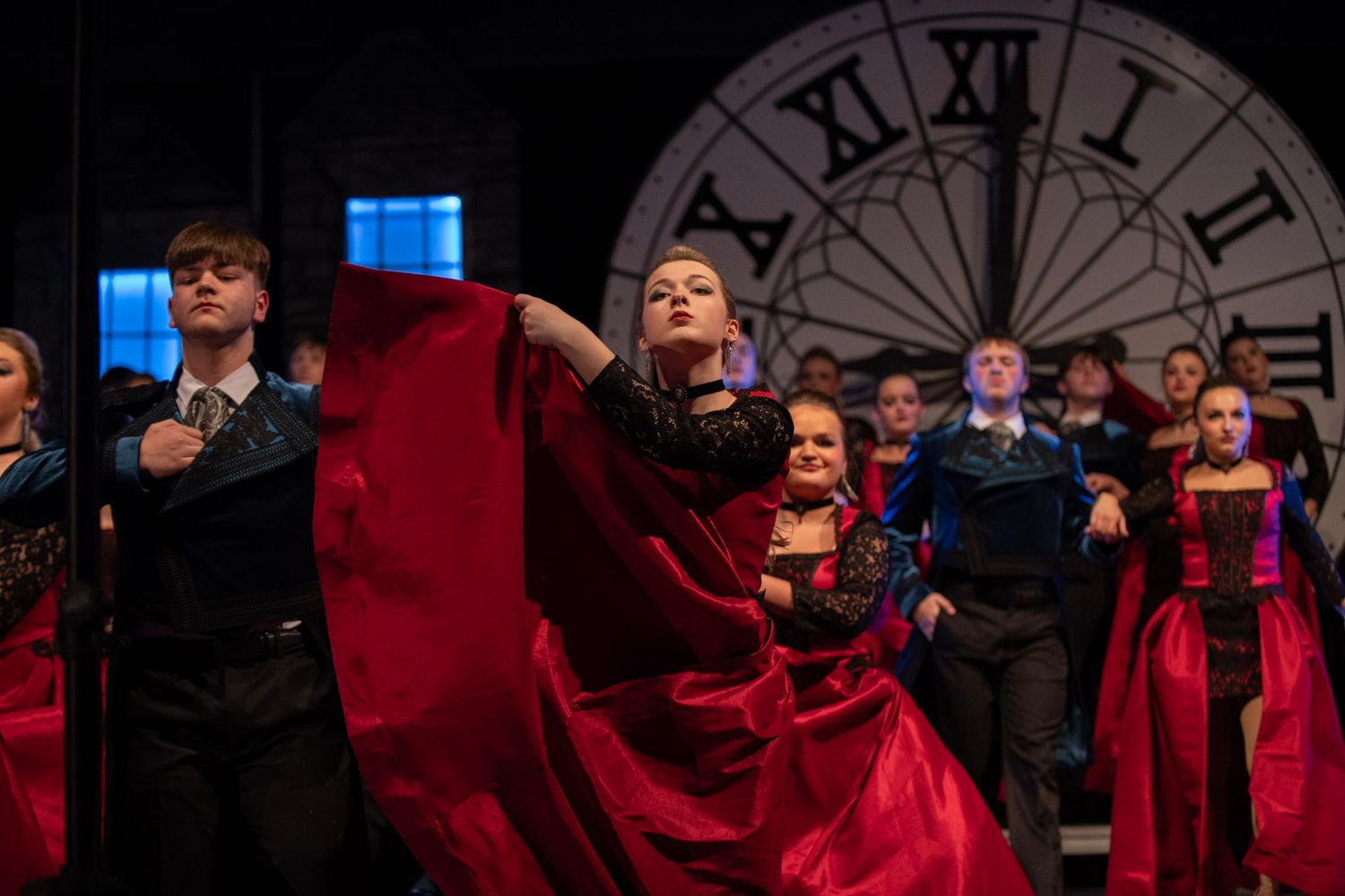 Dancers in red dresses performing on stage with a large clock backdrop.