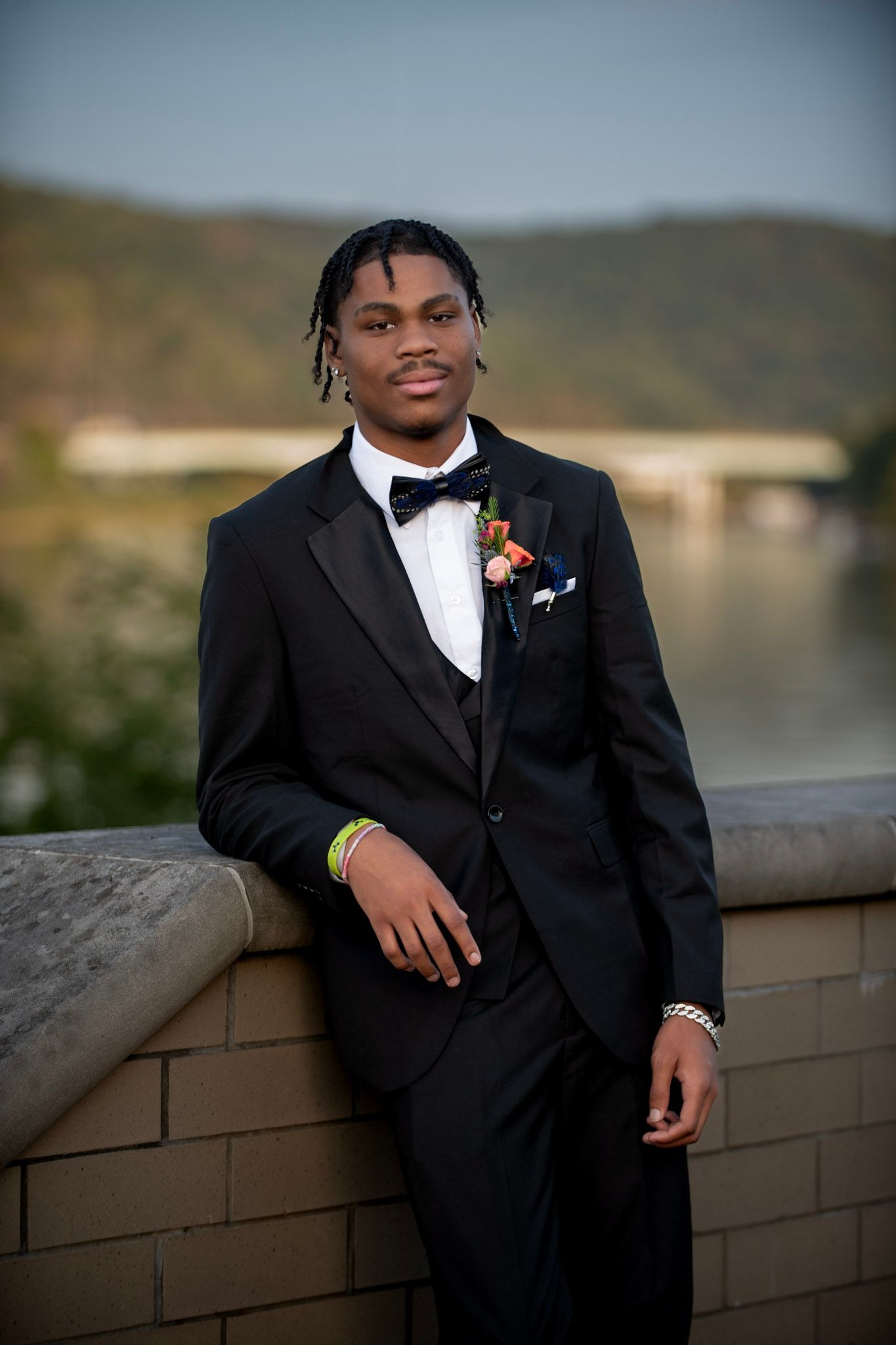 Person in formal black tuxedo poses against stone wall during sunset.