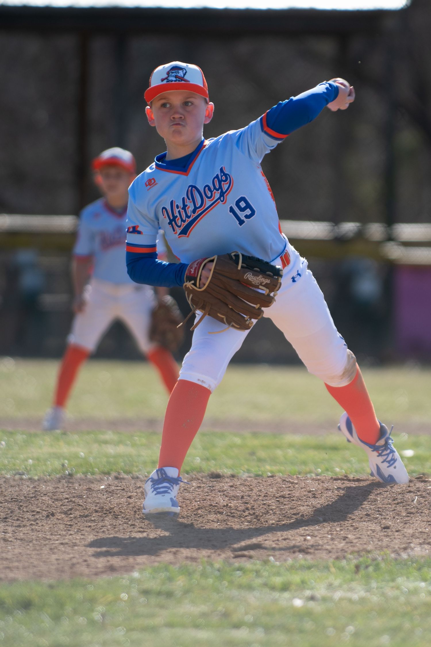 Youth baseball pitcher in blue uniform winds up to throw from the mound.