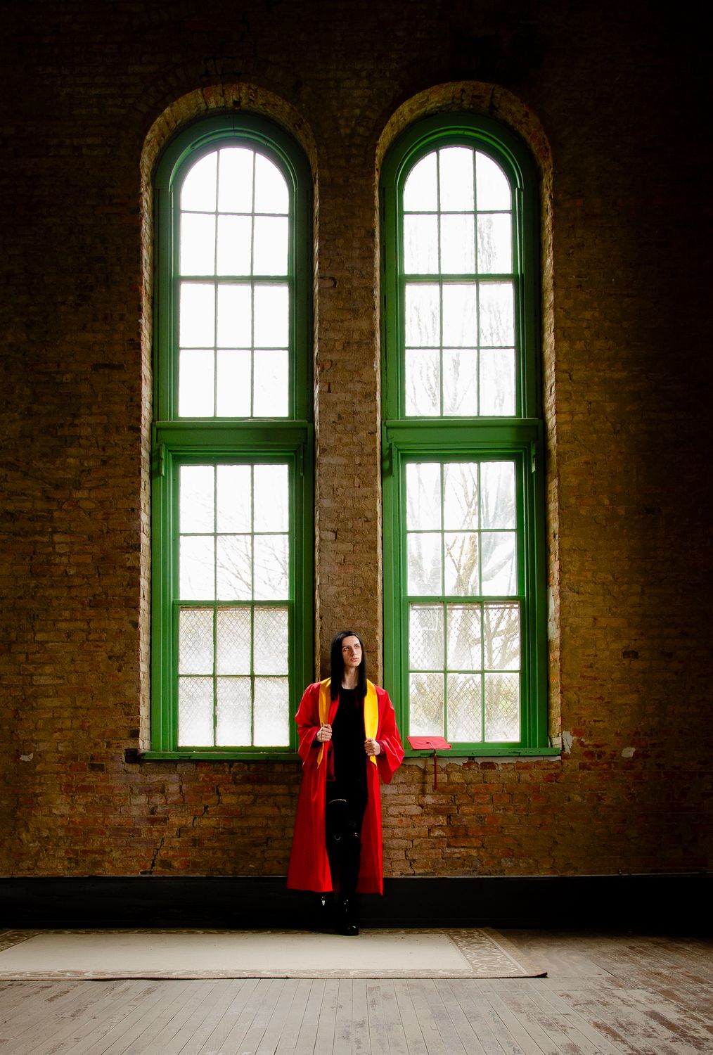 Figure in a striking red outfit stands between two tall arched green windows in a rustic brick interior space.