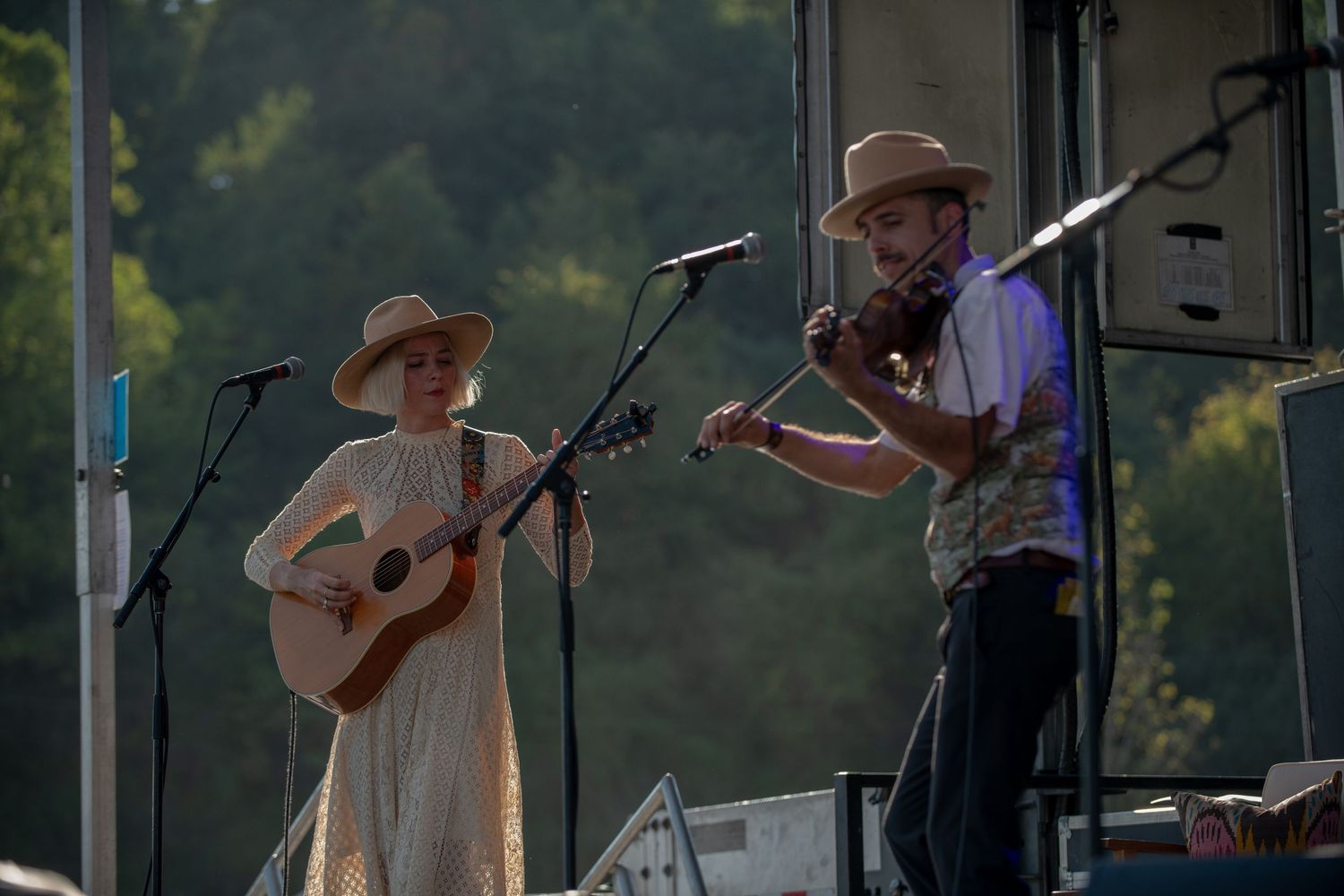 Musicians perform with acoustic guitars and instruments on an outdoor stage at dusk.