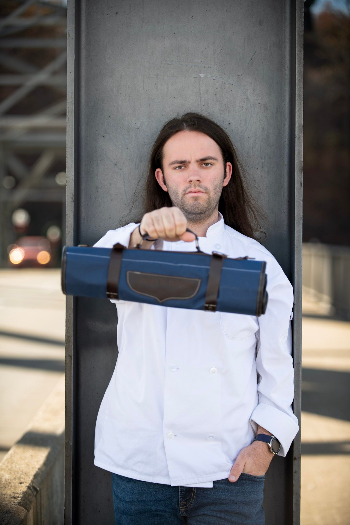 Person in white shirt holding blue knife roll bag against industrial background.