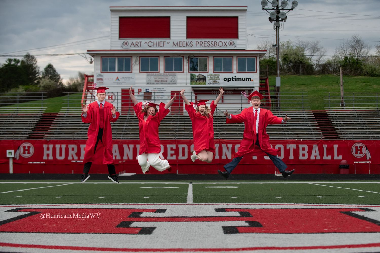 Group of graduates in red gowns jumping in celebration on football field with stadium seats behind.