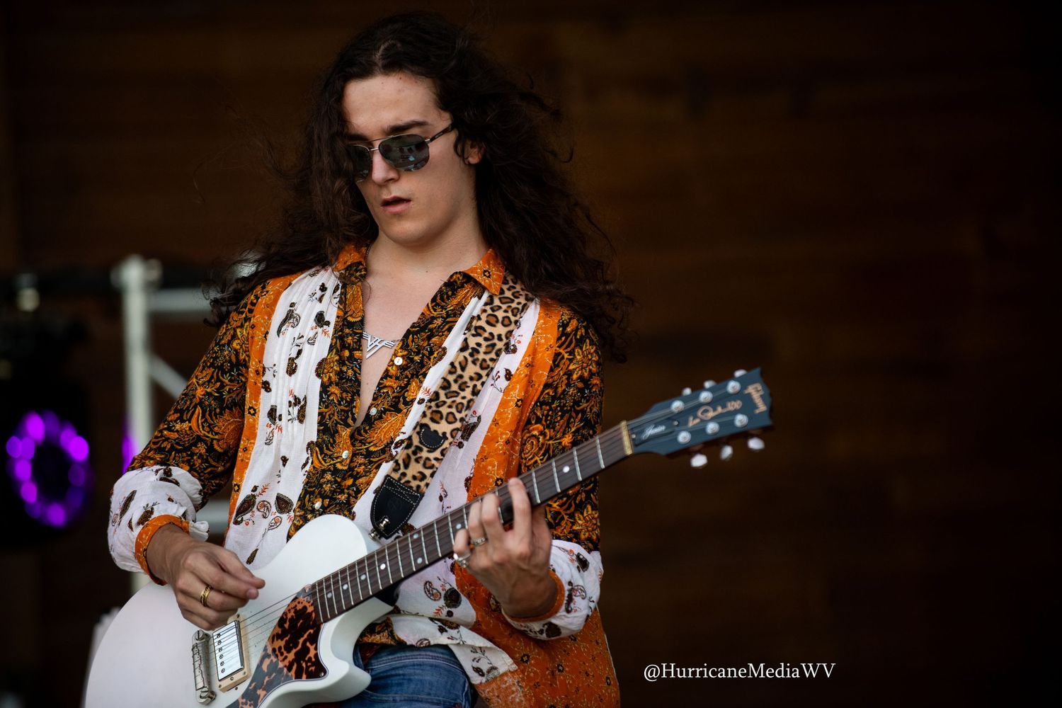 A musician performs with an acoustic guitar wearing sunglasses and a leopard print top at an outdoor show.