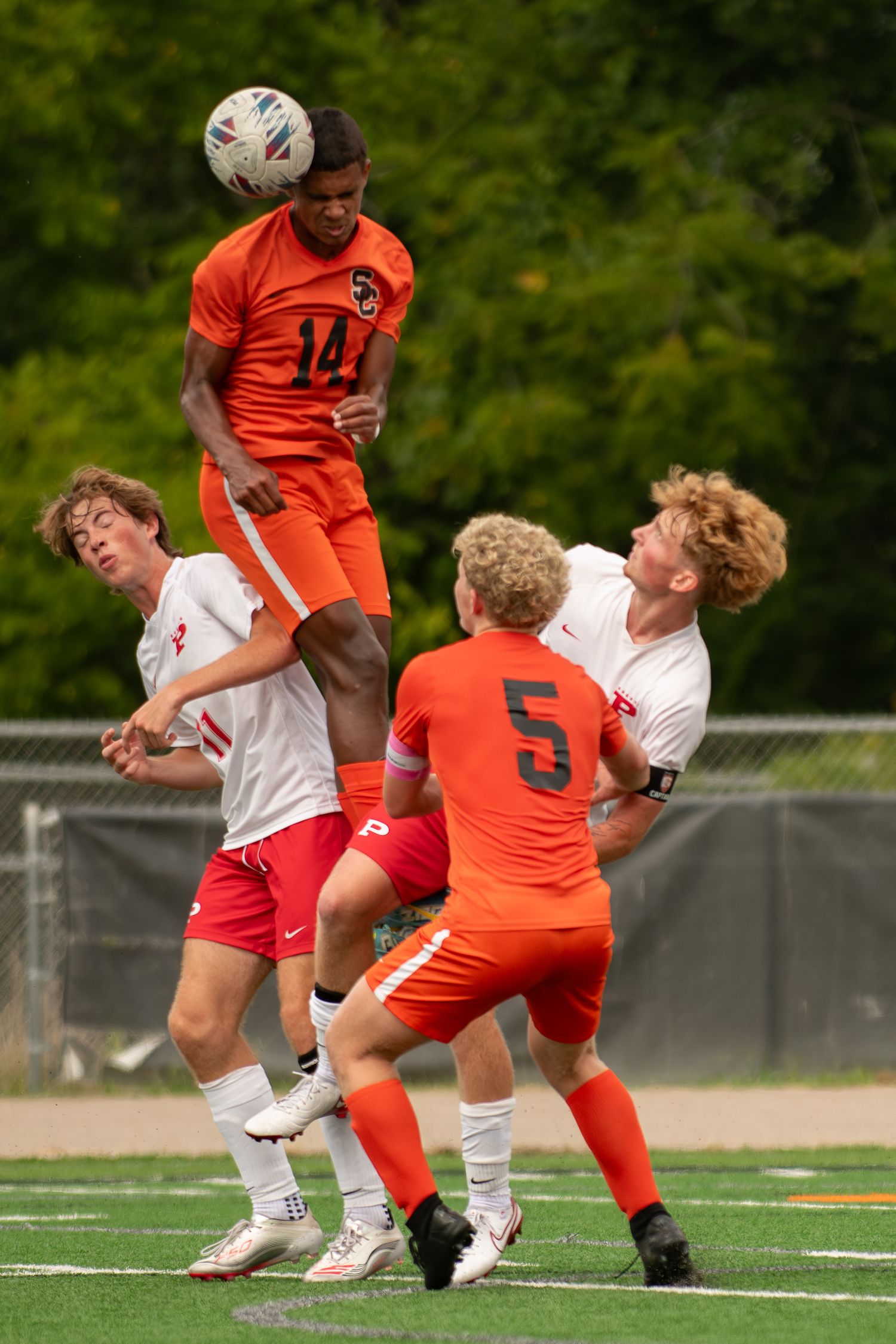 Soccer players collide during an intense match between teams in orange and white uniforms on an outdoor field.