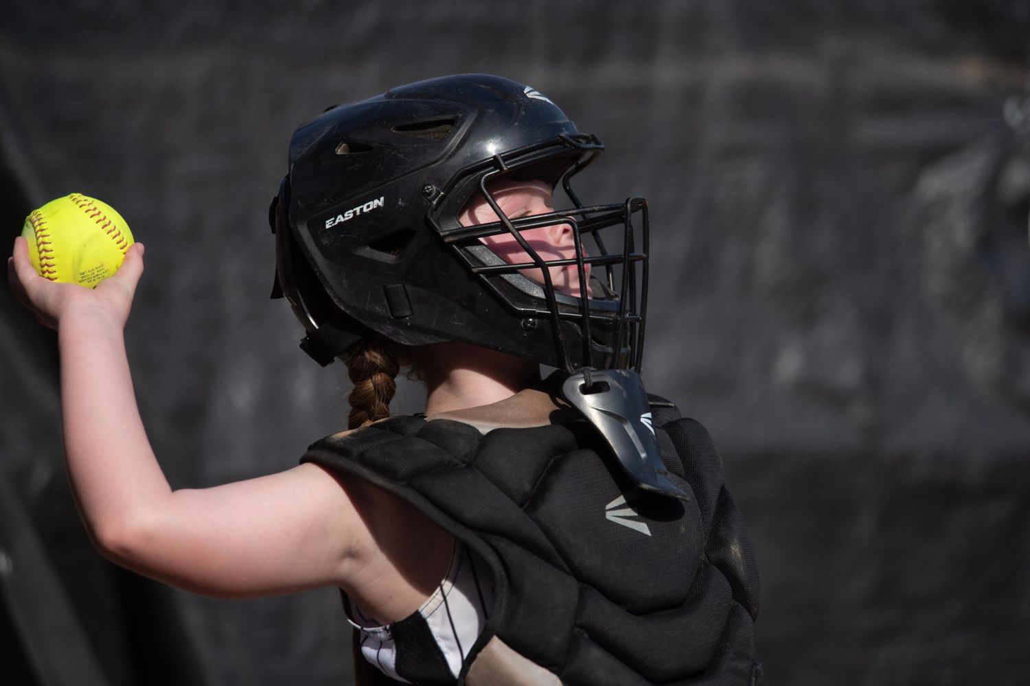 Softball player in black protective gear and helmet preparing to throw yellow ball during game.