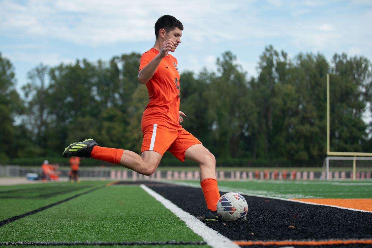 Soccer player in bright orange uniform kicks ball on artificial turf field with trees in background.