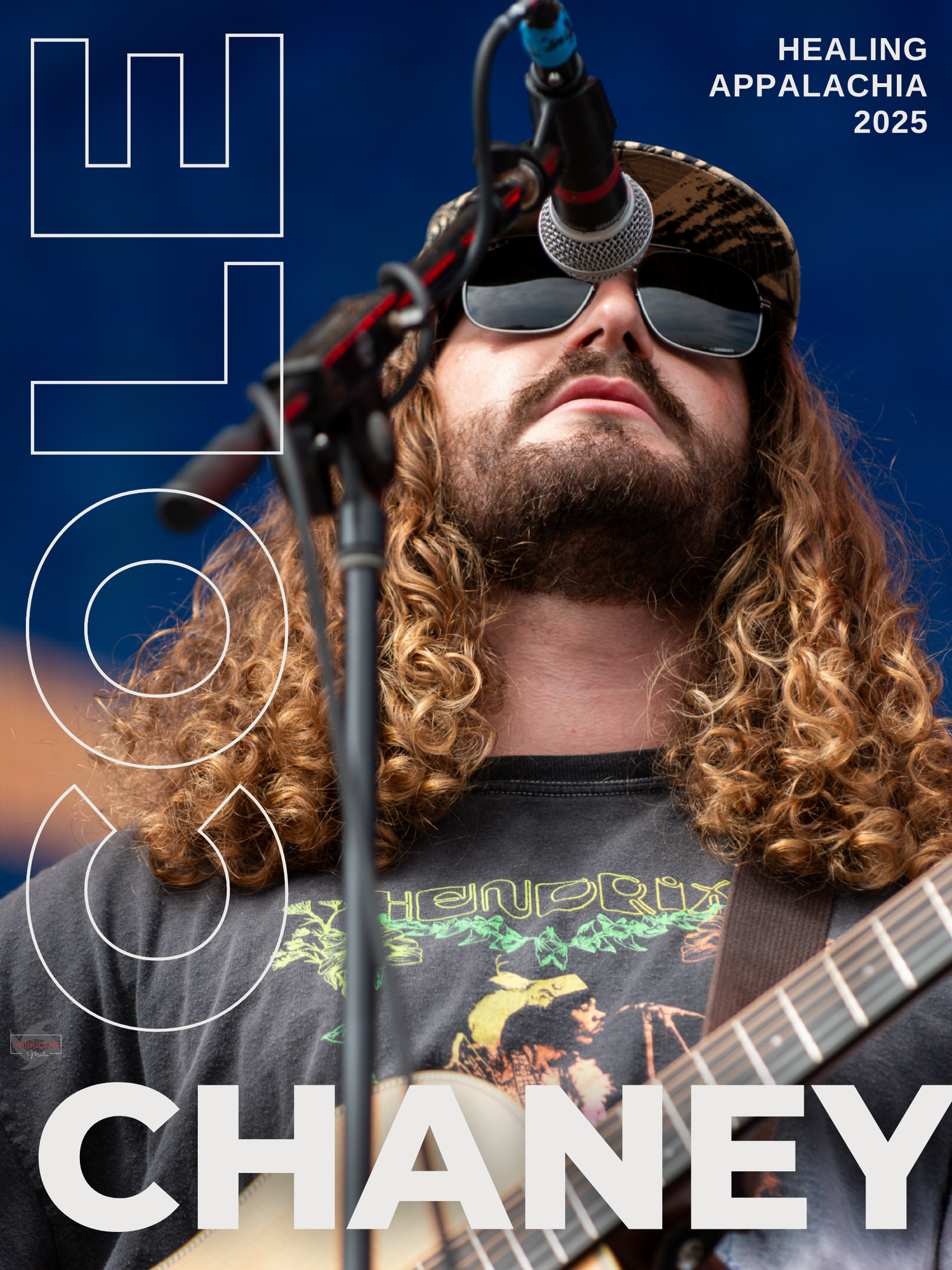 A musician with curly hair and sunglasses performs on stage with a guitar during a concert performance.