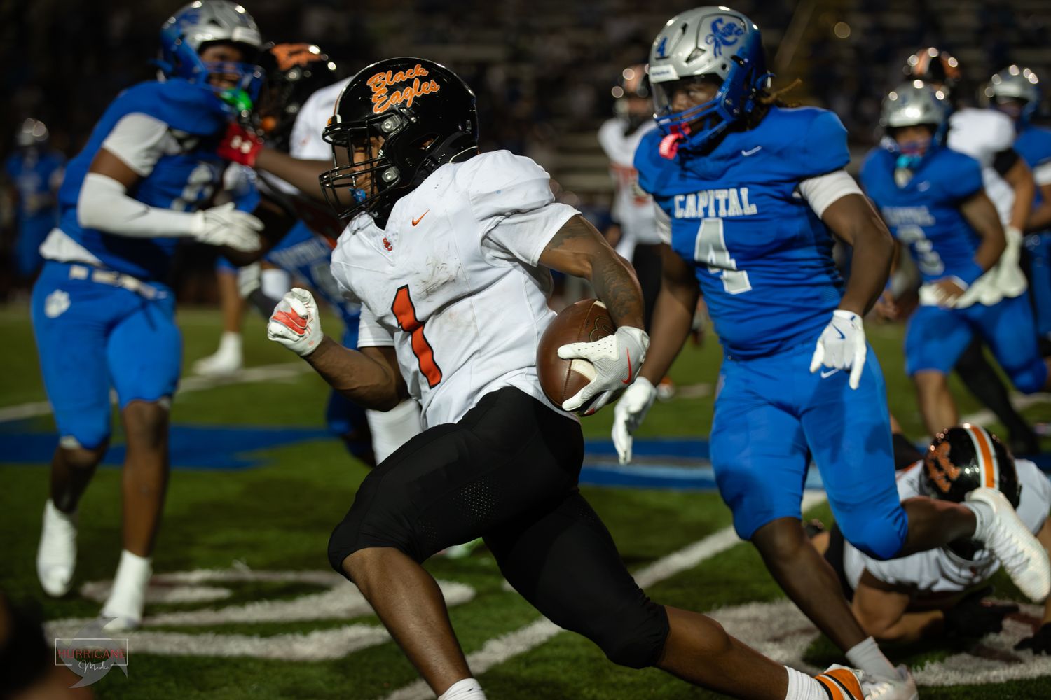 Football players engage in an intense play during a night game with blue and white uniforms visible on the field.