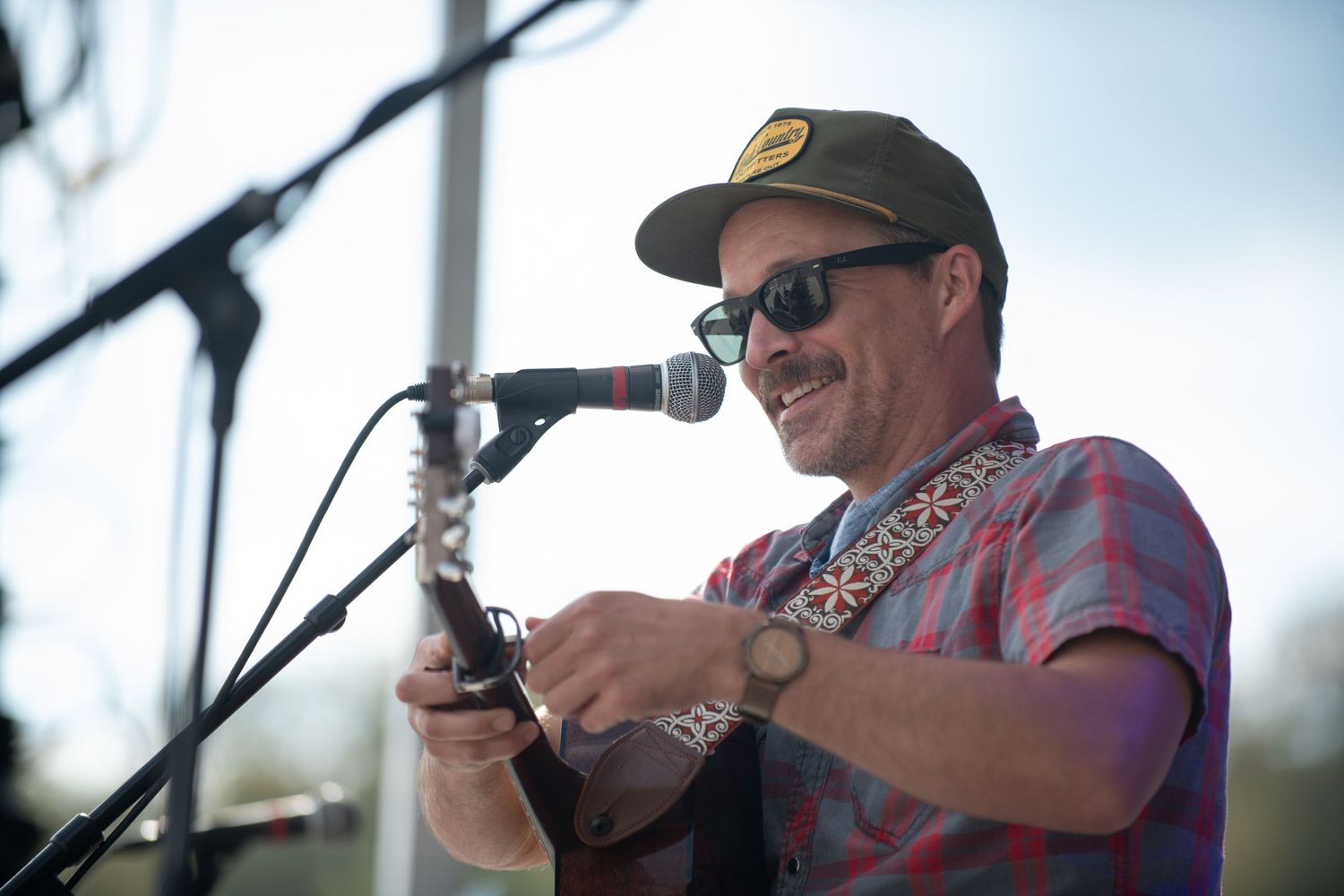 A performer in sunglasses and baseball cap smiles while speaking into microphone during outdoor performance.