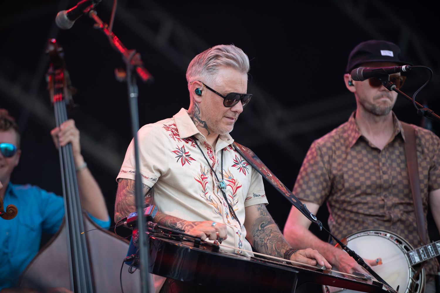 Musicians perform on stage at an outdoor concert with one playing upright bass and wearing sunglasses and a white shirt.