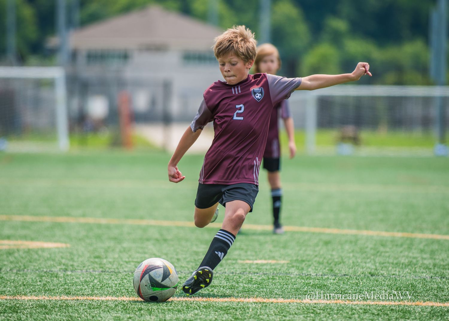 Young soccer player in maroon jersey kicking ball on green turf field.