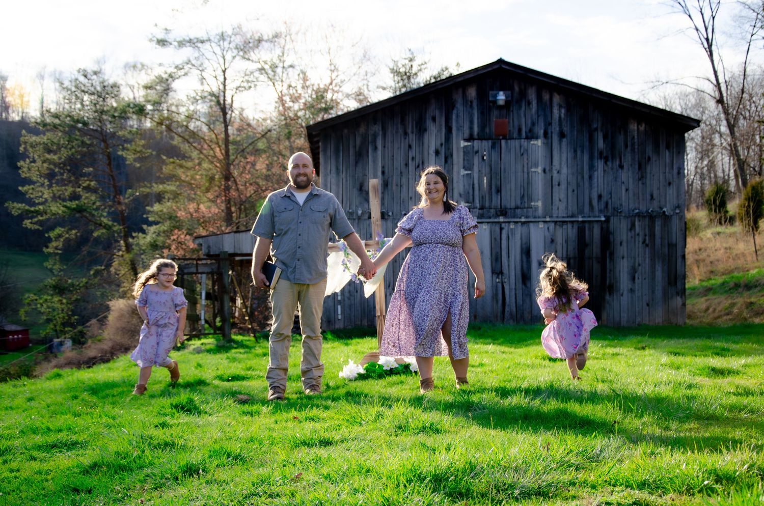 Family enjoying a playful moment on green grass in front of a rustic barn during golden hour.