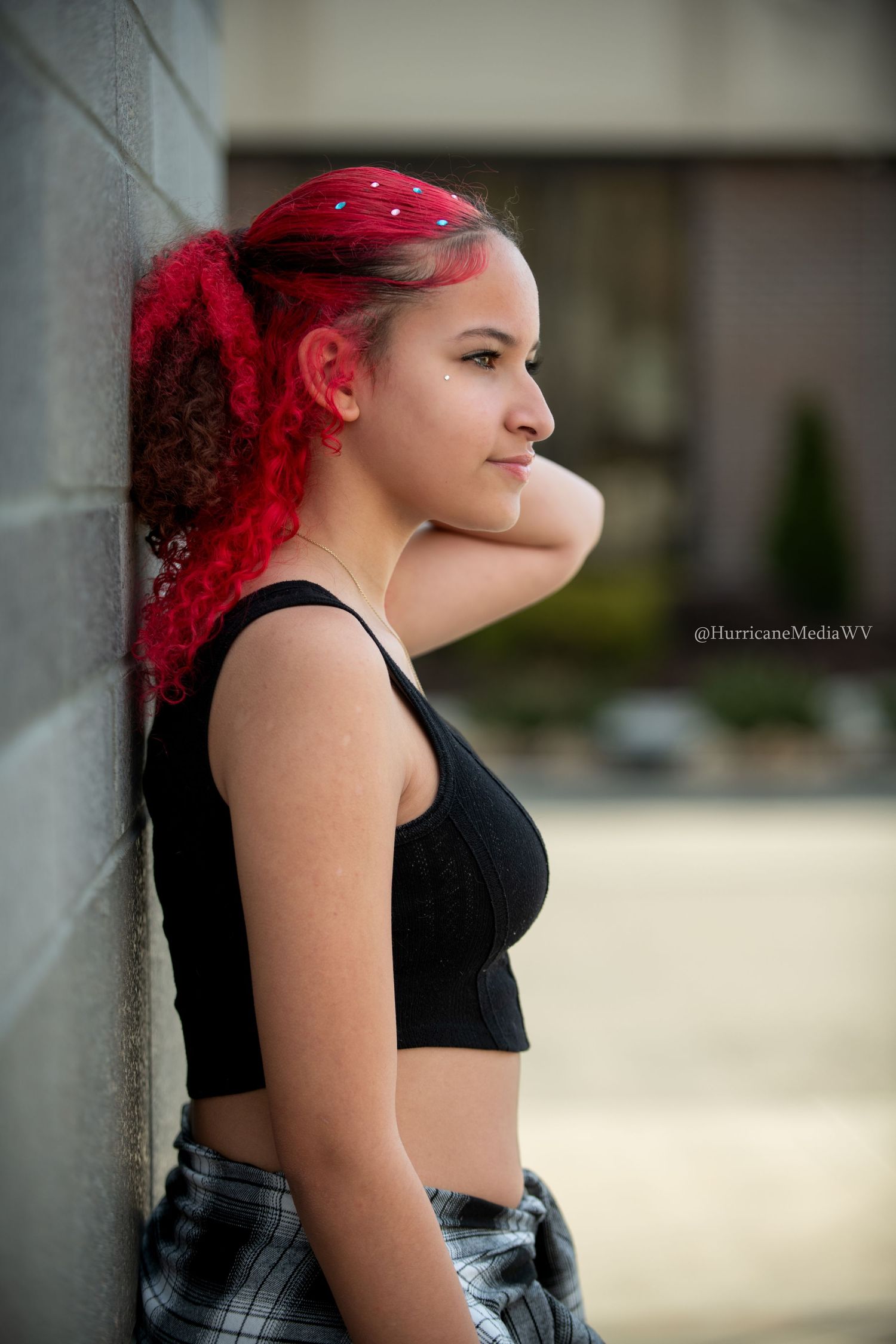 A person with bright red hair wears a black tank top and plaid skirt while posing outdoors against a white wall.