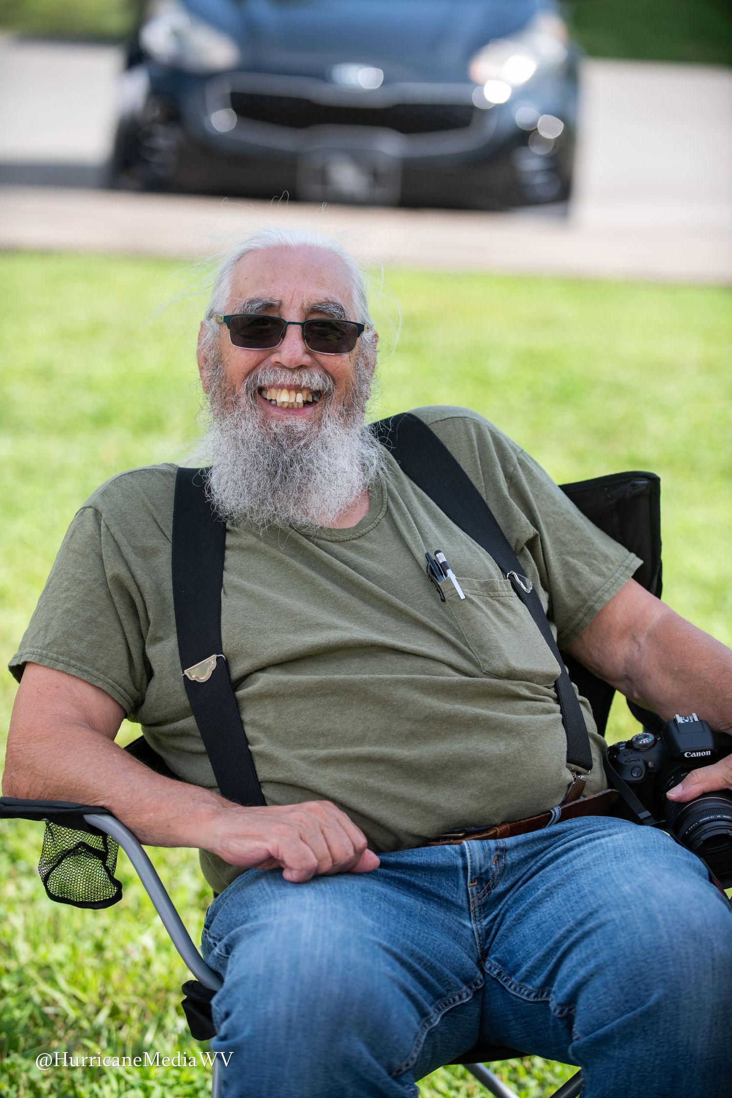 A cheerful photographer with a long gray beard wearing sunglasses and suspenders sits outdoors with a car in background.