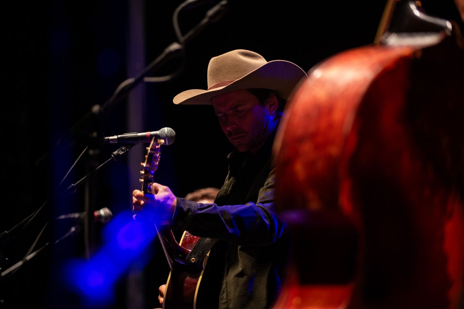 A performer plays guitar on a dimly lit stage with blue lighting effects in the background.