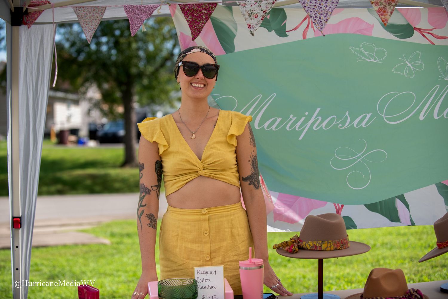 Vendor wearing yellow summer outfit and sunglasses stands at outdoor market booth with decorative bunting overhead.