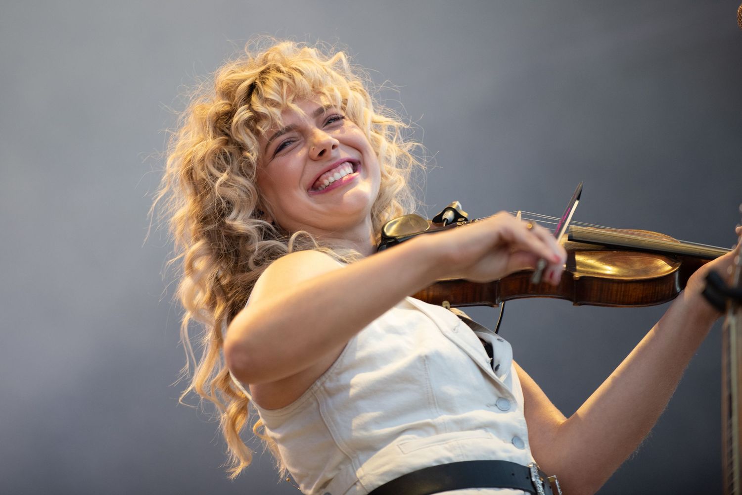 Musician plays violin with joyful expression while performing on stage in white outfit against gray background.