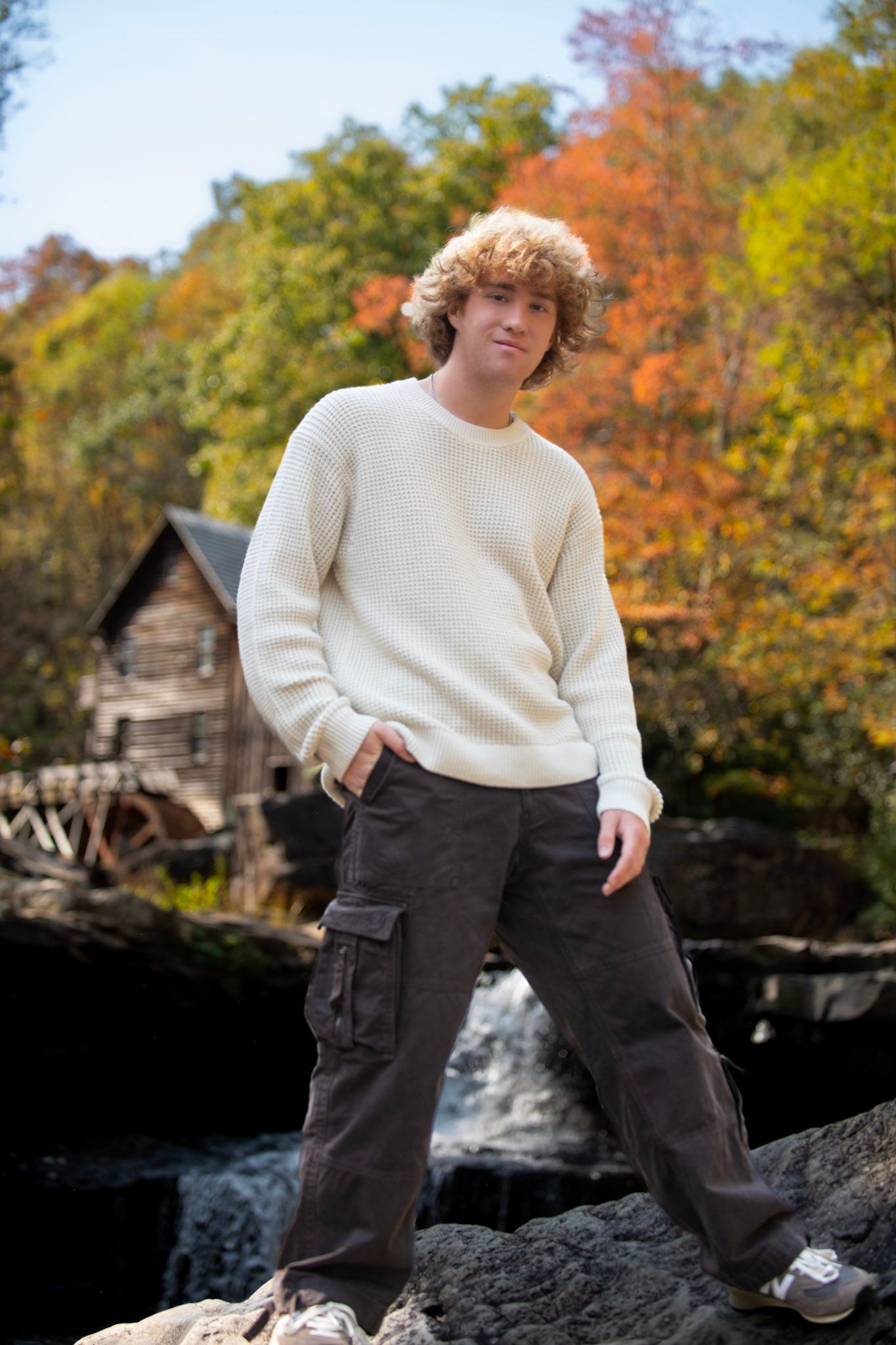 A person in a cream sweater poses on rocks near a waterfall with autumn foliage in background.