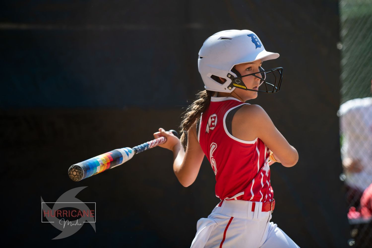 Softball player in red jersey and white helmet swings bat during game with braided hair visible from helmet.