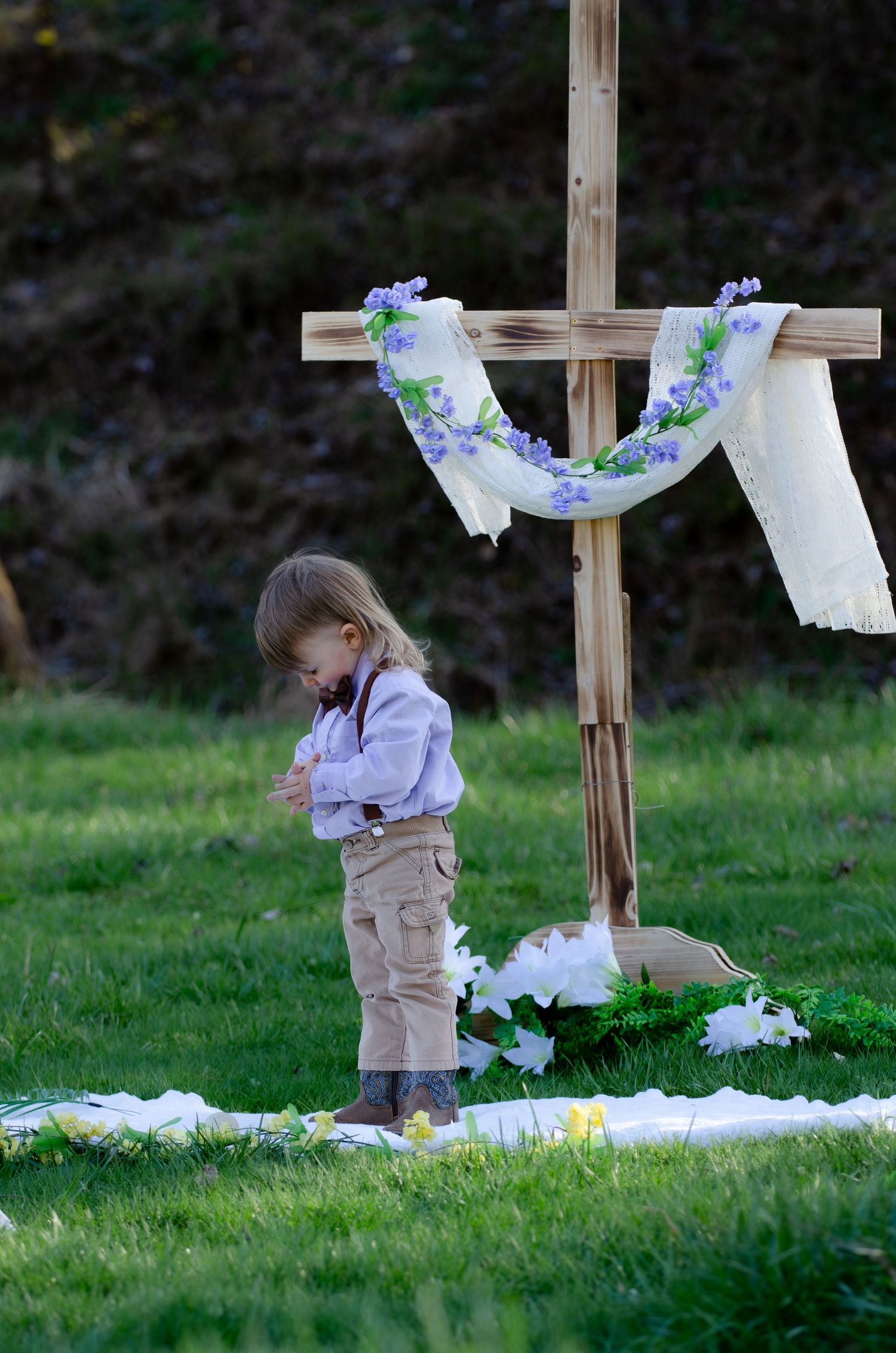 Young child stands near decorated Easter cross display in grassy field.
