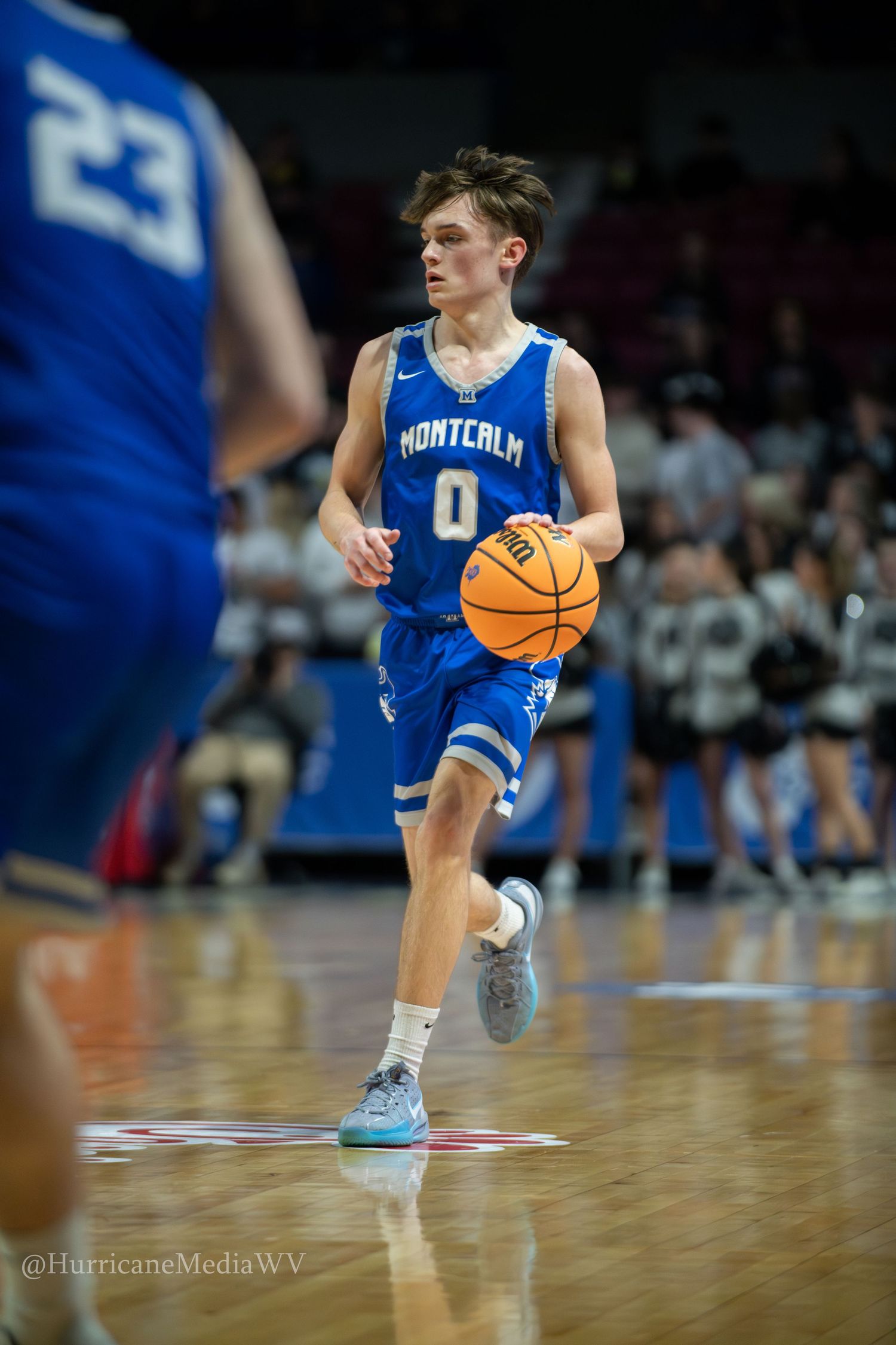 Basketball player in blue and white uniform dribbles the ball up court during game action.