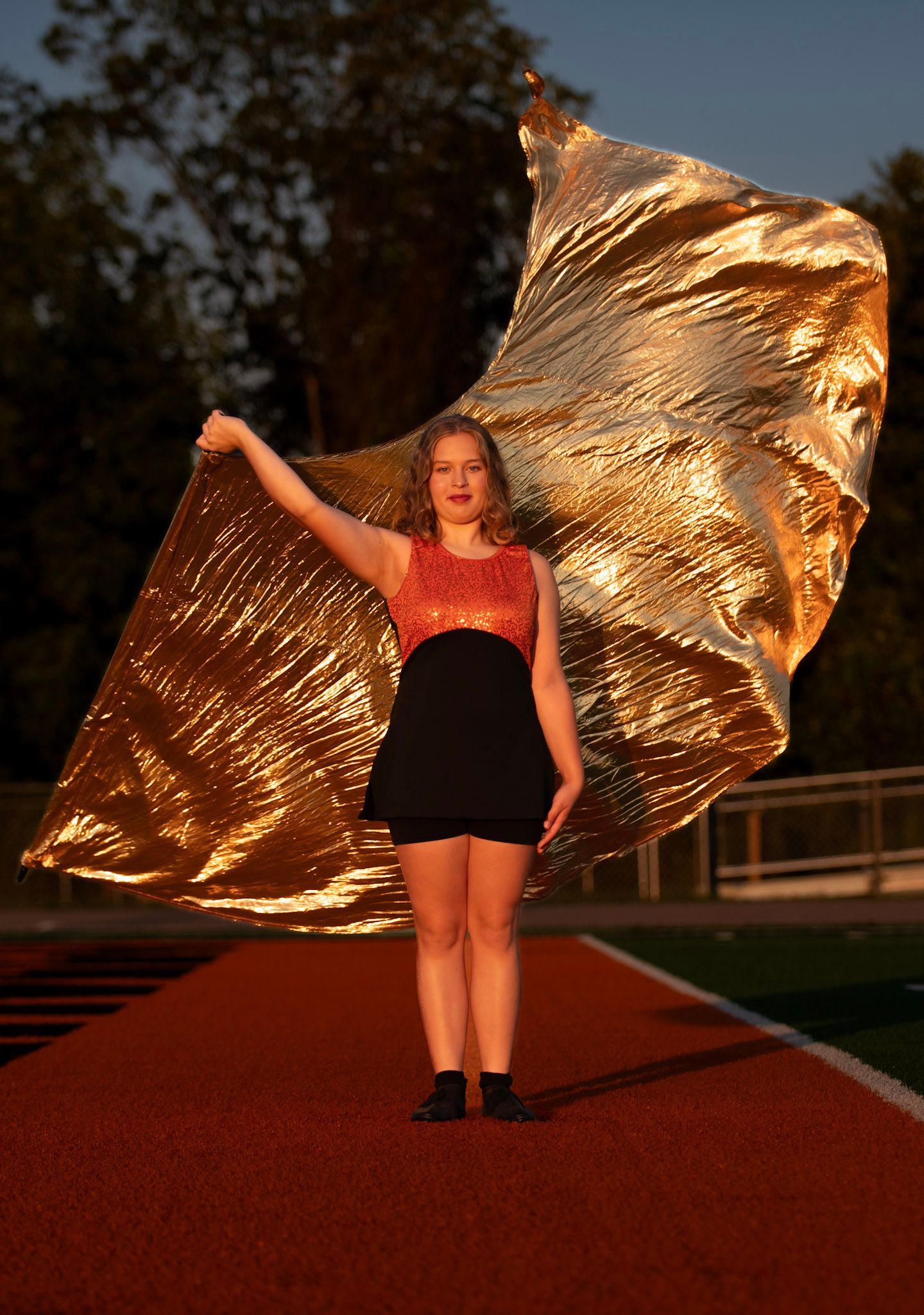 Color guard performer with golden flag practices routine on red track at sunset.