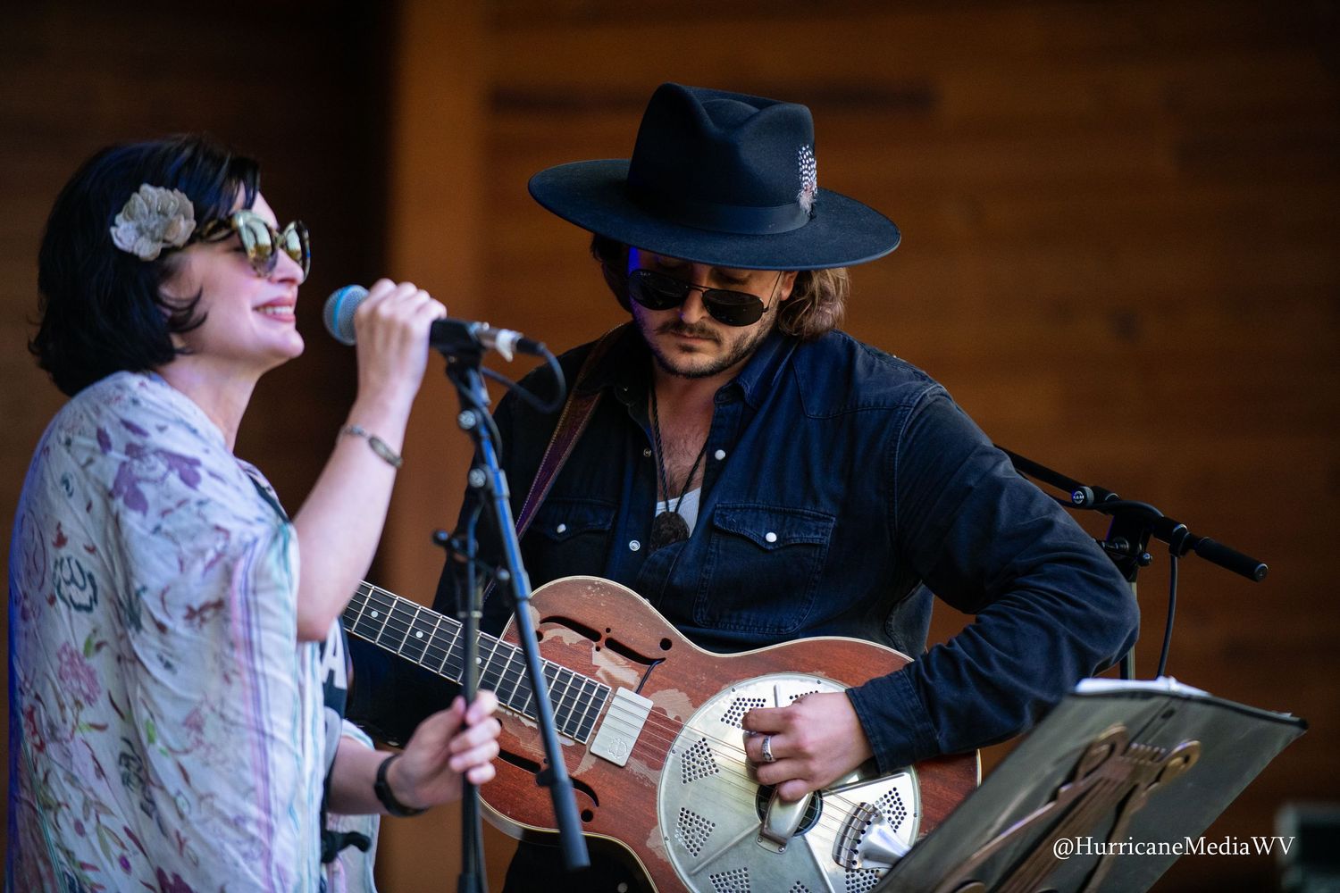 Musicians perform together on stage during a live concert with one wearing a black hat and sunglasses.