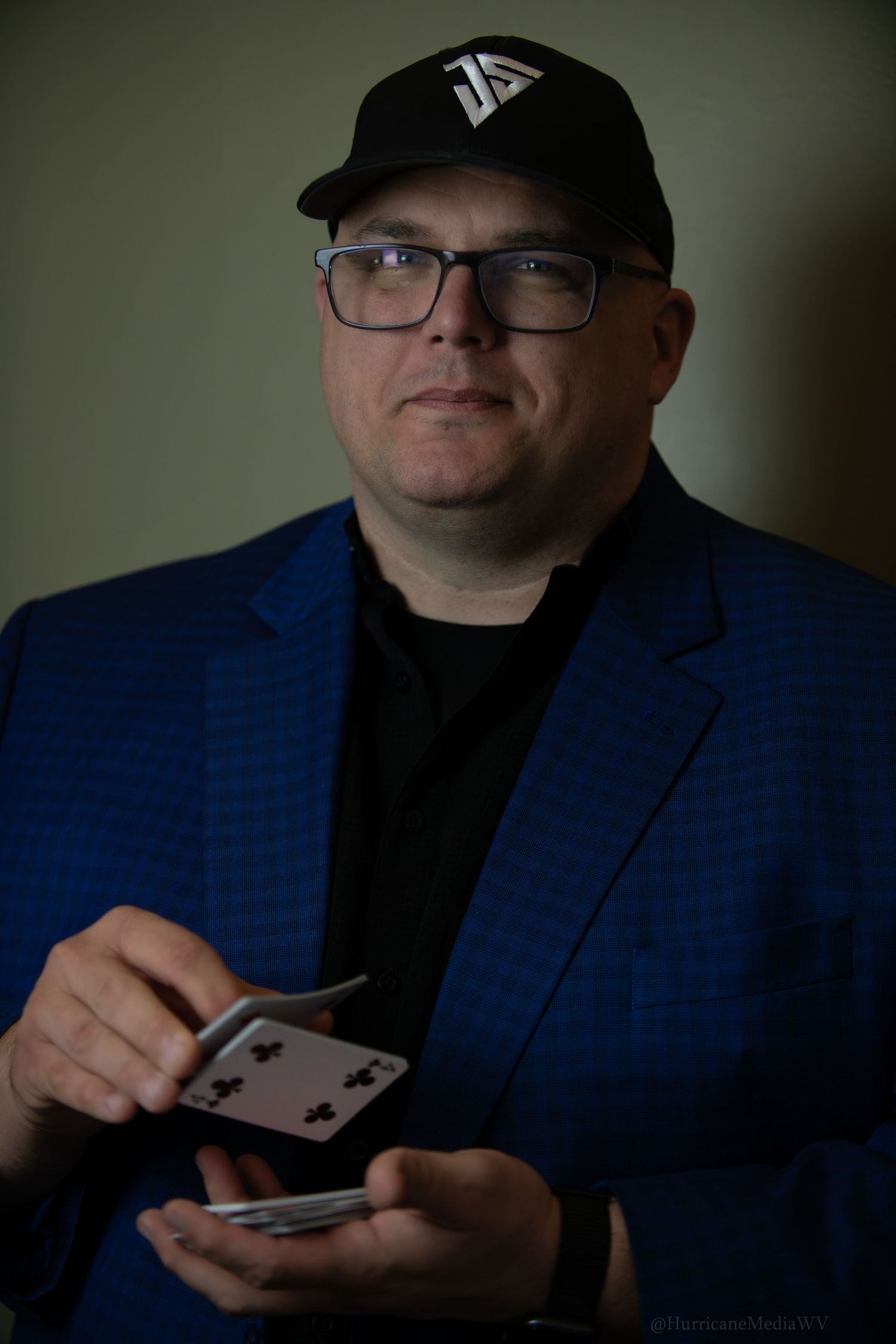 Person in blue blazer and baseball cap holding playing cards in dramatic lighting.