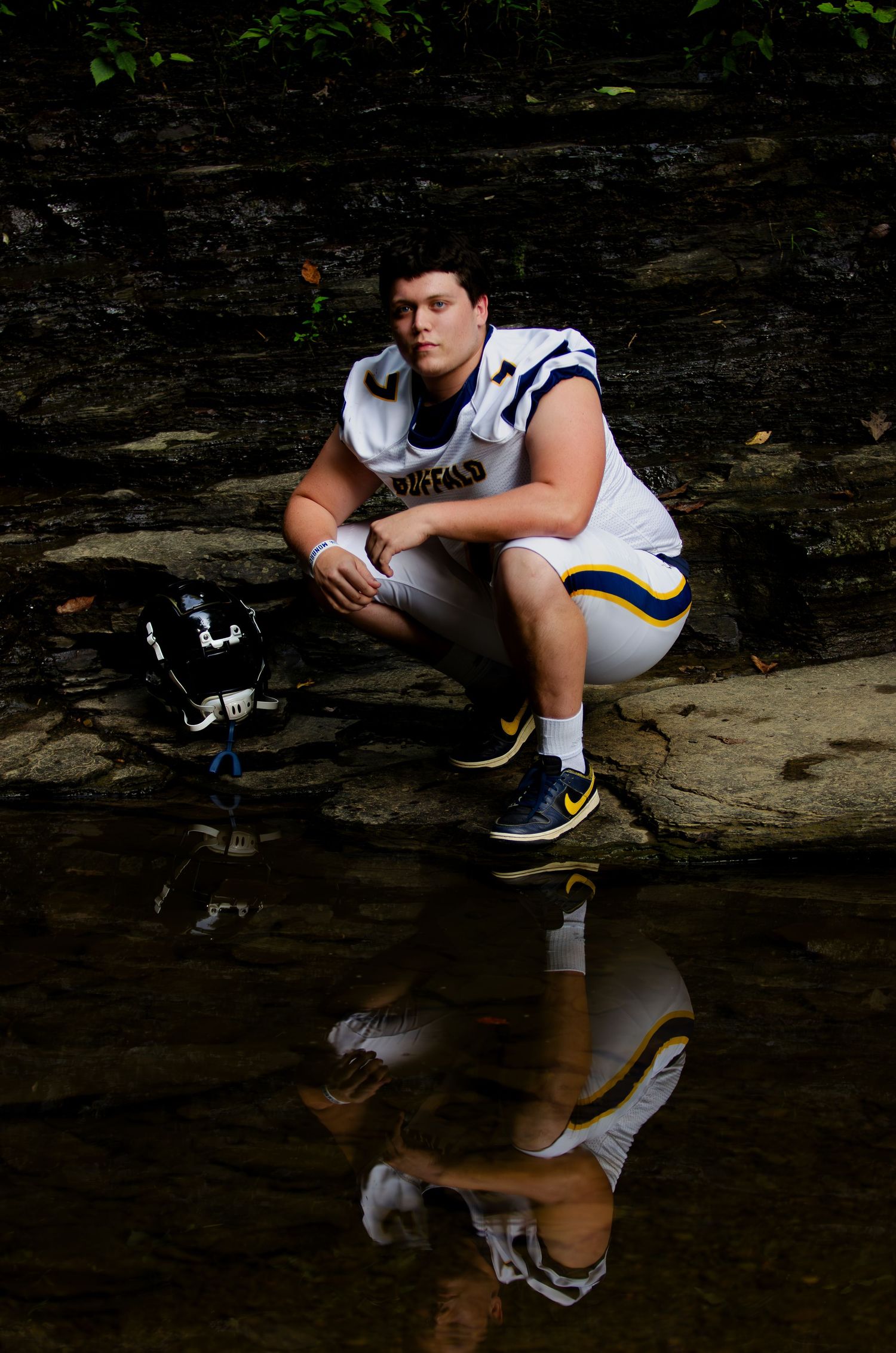 Football player in white uniform sits reflectively by water's edge in outdoor setting.