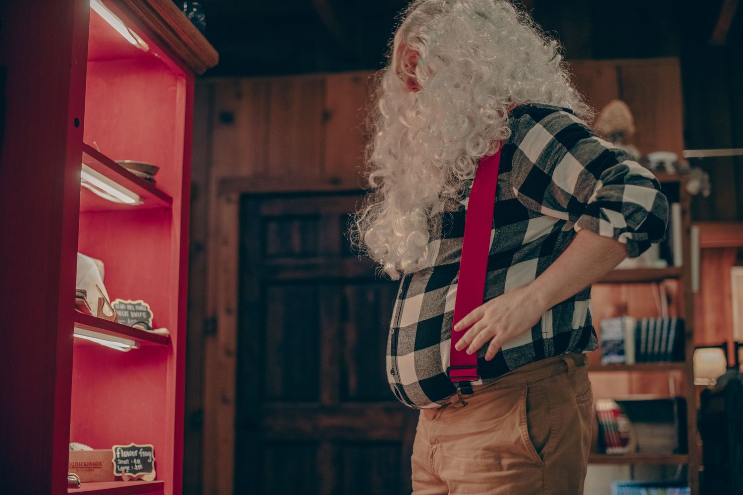 Person wearing plaid shirt and overalls works on red cabinet in rustic workshop setting.