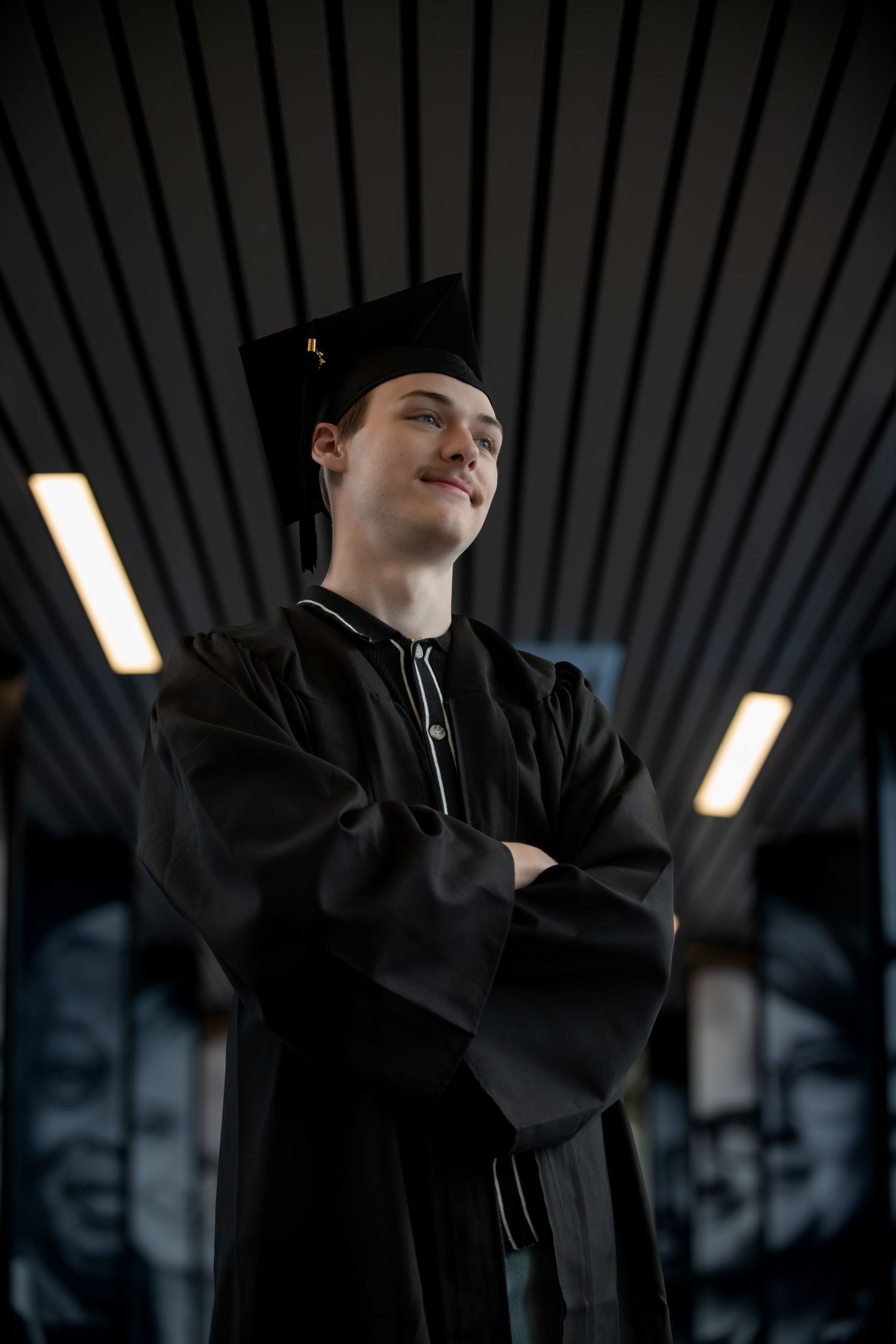 Graduate in traditional black cap and gown standing with crossed arms.