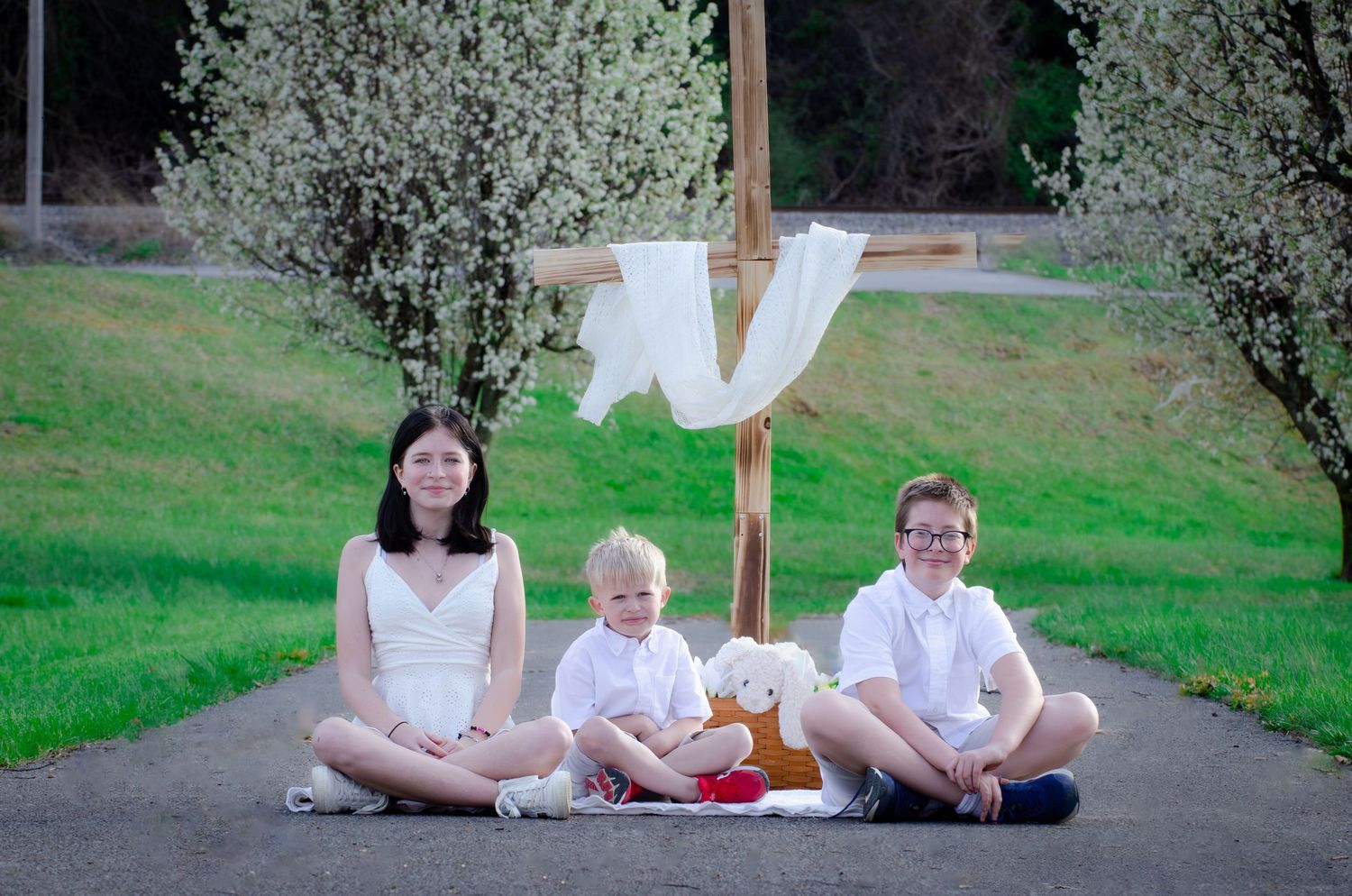 Three people sitting on pathway with stuffed bear under hanging white fabric and blooming trees.