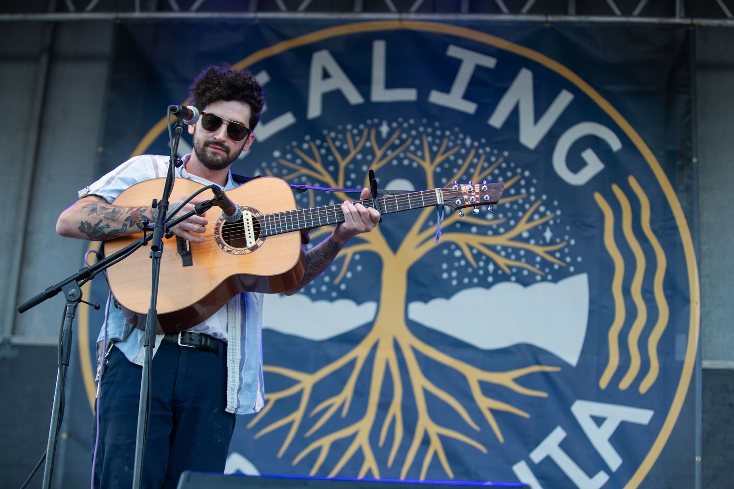 Musicians perform on acoustic guitars at an outdoor festival with a blue circular logo banner in the background.