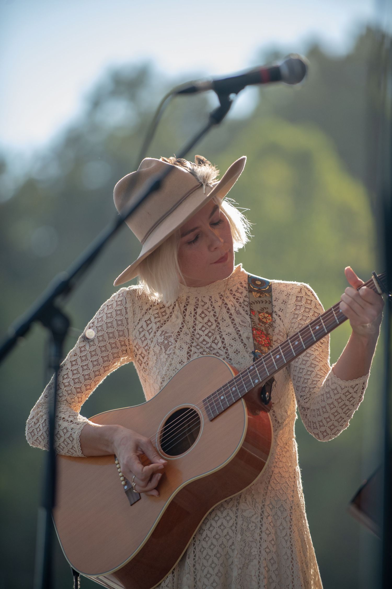 A musician in a white dress and wide-brimmed hat performs with an acoustic guitar on an outdoor stage in natural lighting.