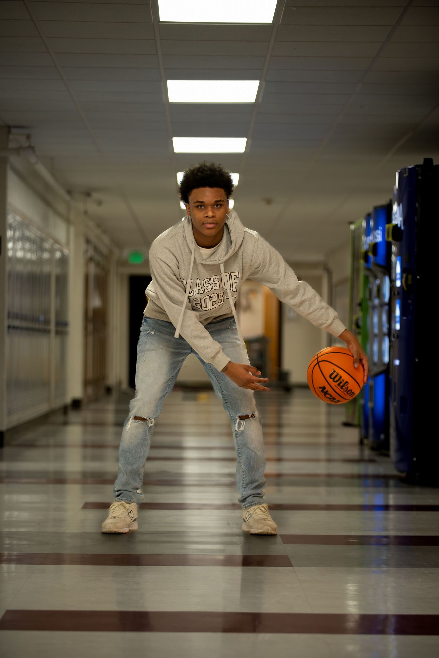 Basketball player in ripped jeans and gray sweatshirt dribbling in school hallway with lockers and fluorescent lights.