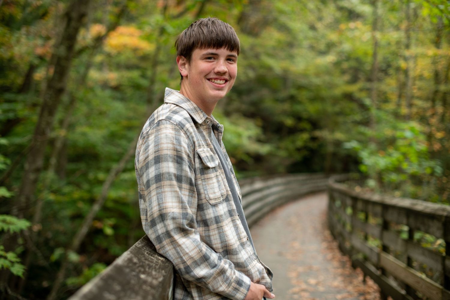 A person in a plaid shirt walks along a wooden boardwalk trail through a lush green forest.