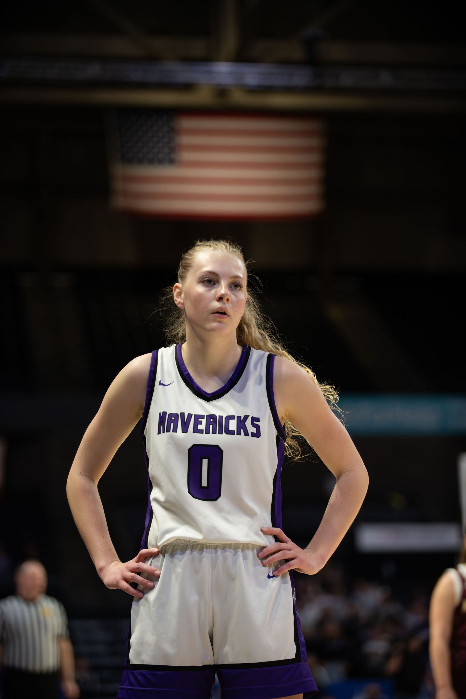 Basketball player in white Mavericks jersey number 0 stands on court during game with American flag visible.