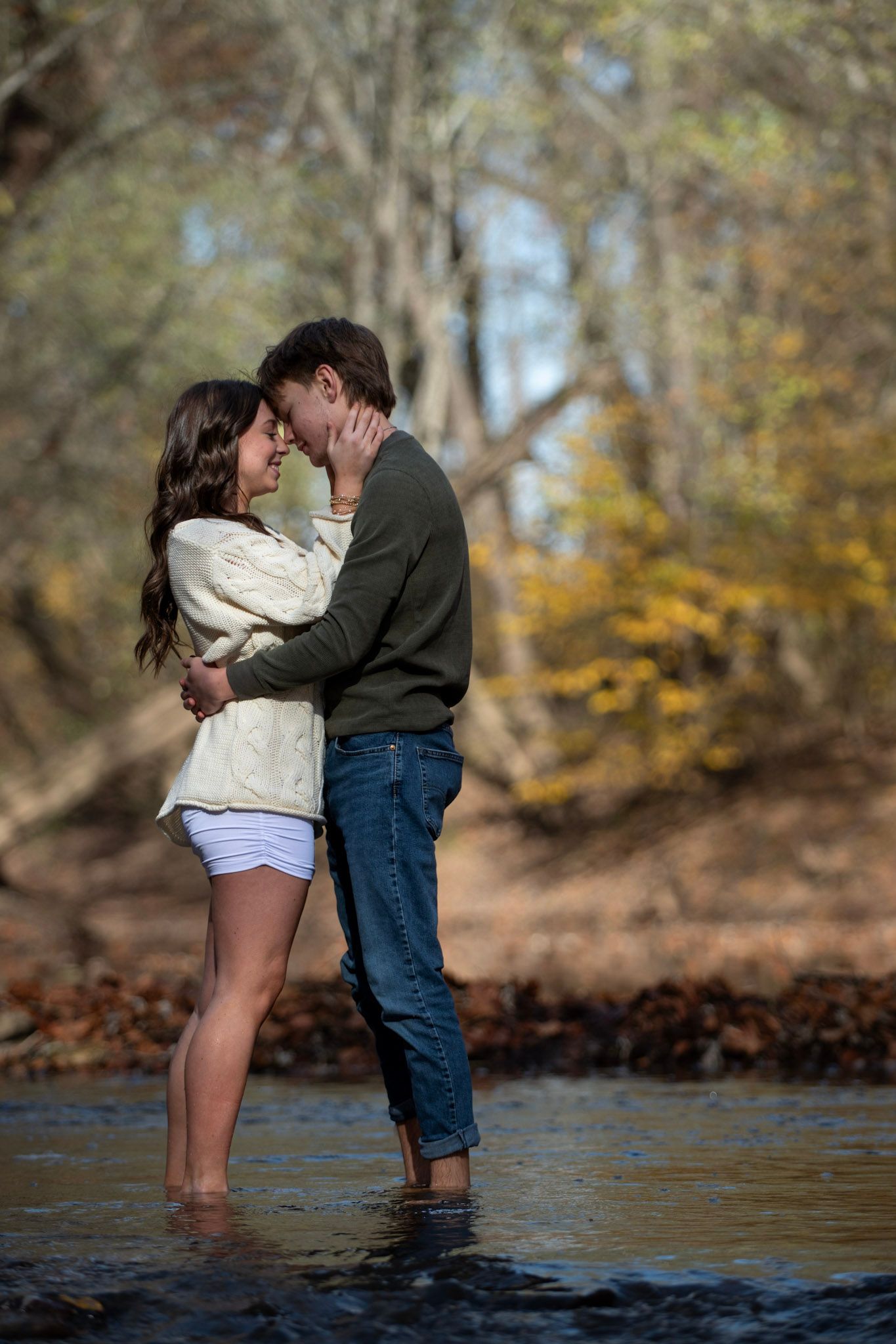 A romantic couple embraces while standing in shallow water during autumn.