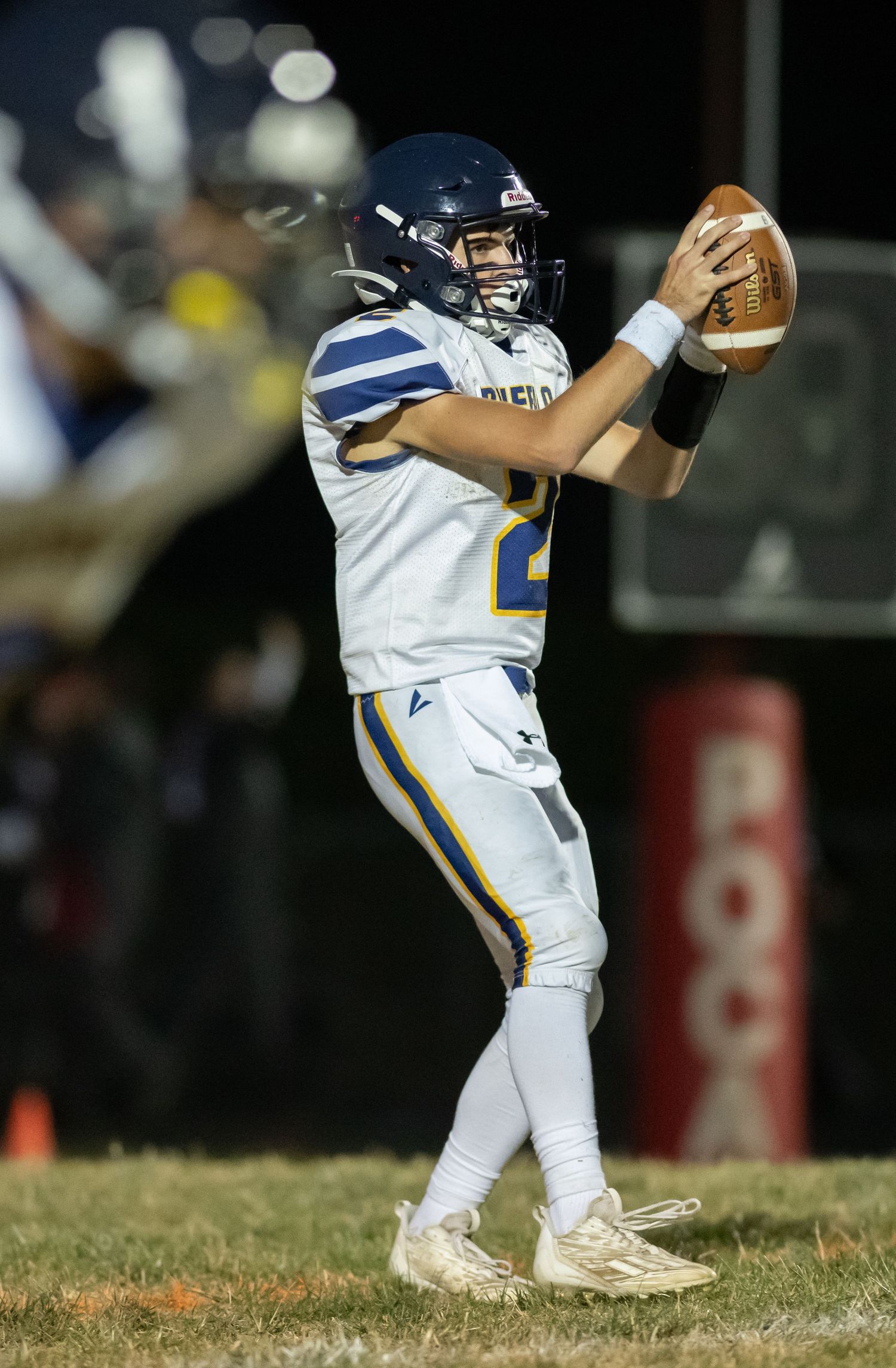 Football quarterback in white and blue uniform prepares to throw the ball during nighttime game.