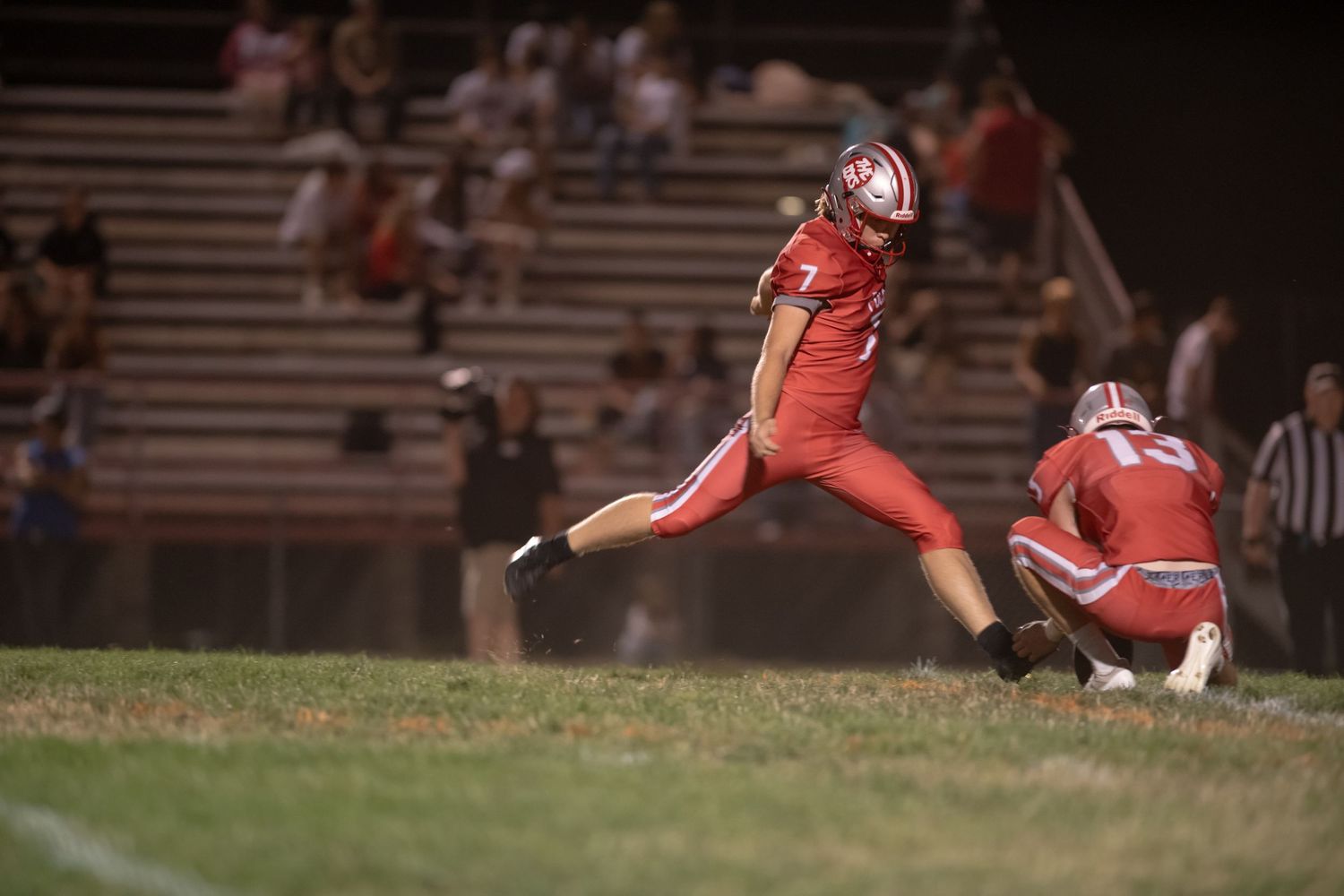 Football kicker in red uniform strikes the ball during a nighttime game while holder kneels on the field.