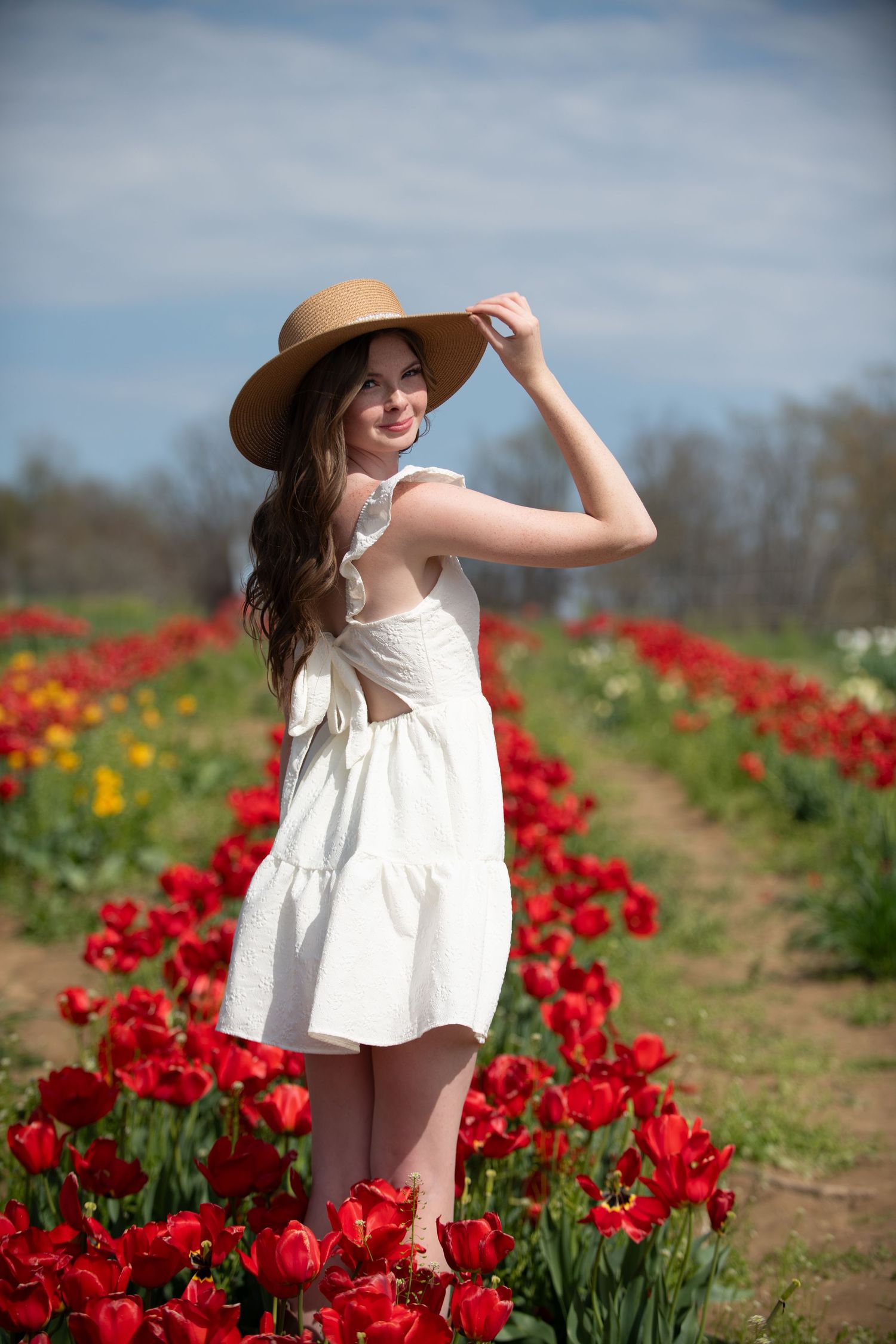 Person in white dress and wide-brim hat poses among rows of red tulips.