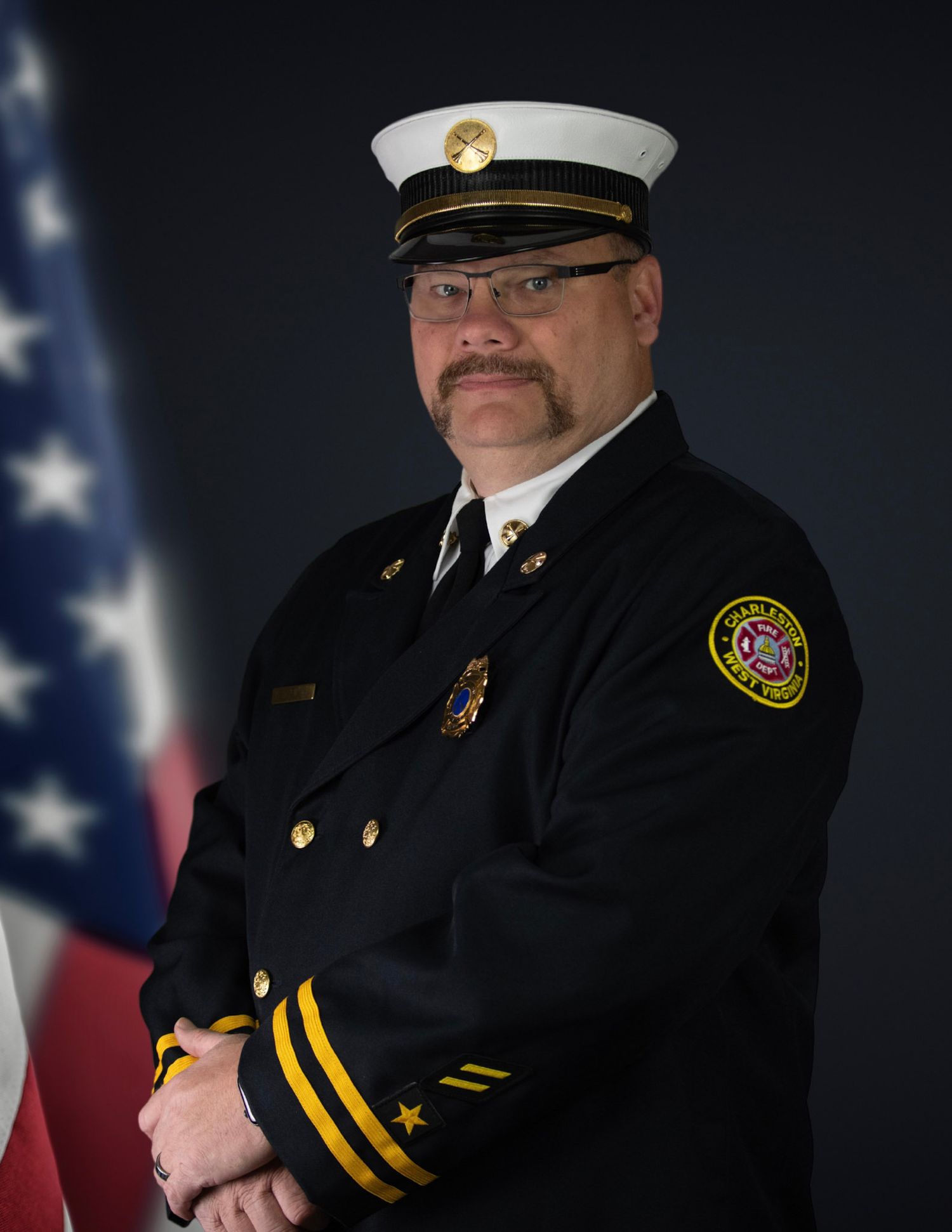 Uniformed officer in formal dress standing at attention with American flag in background.
