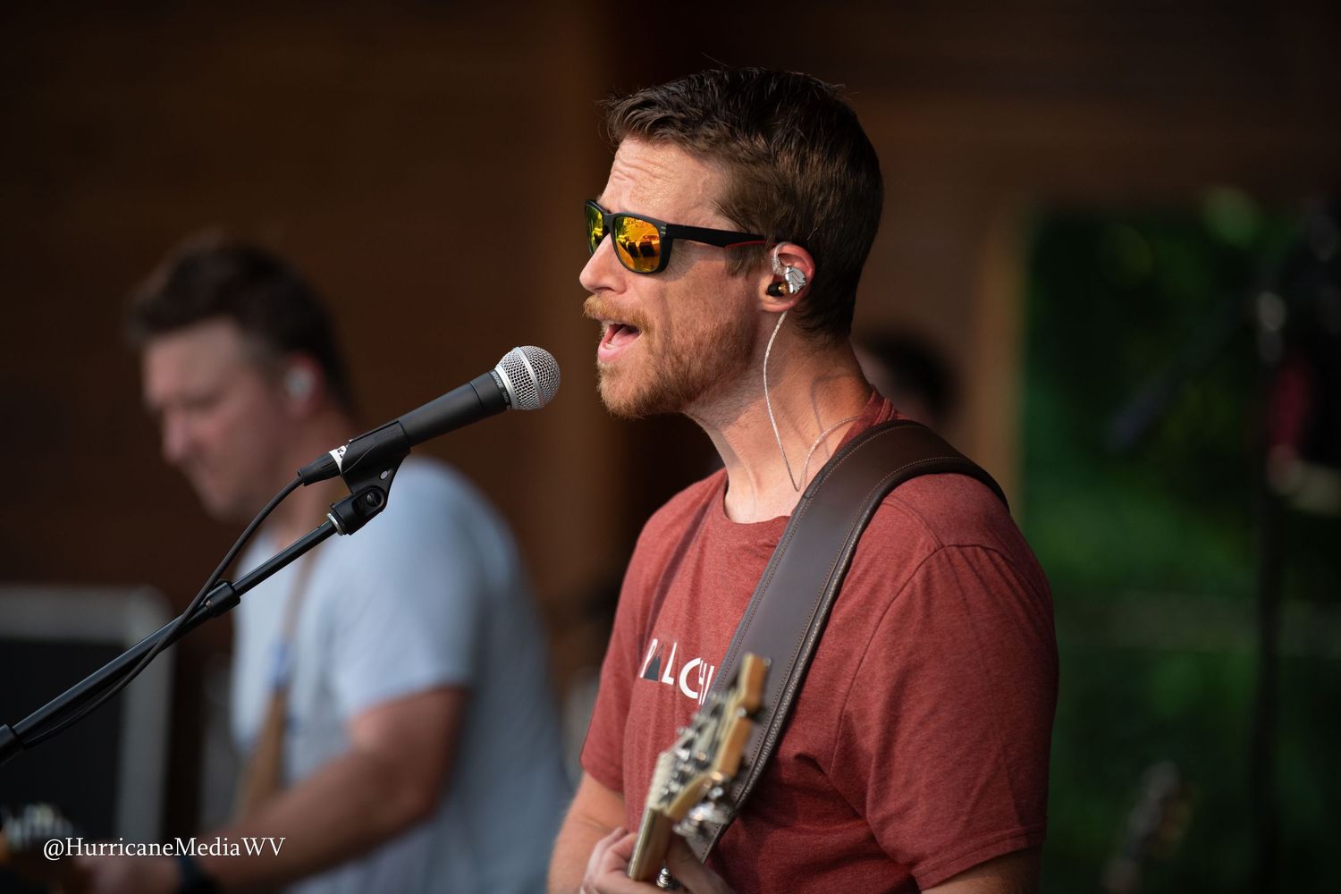 A musician in a red t-shirt performs on stage with sunglasses while singing into a microphone.