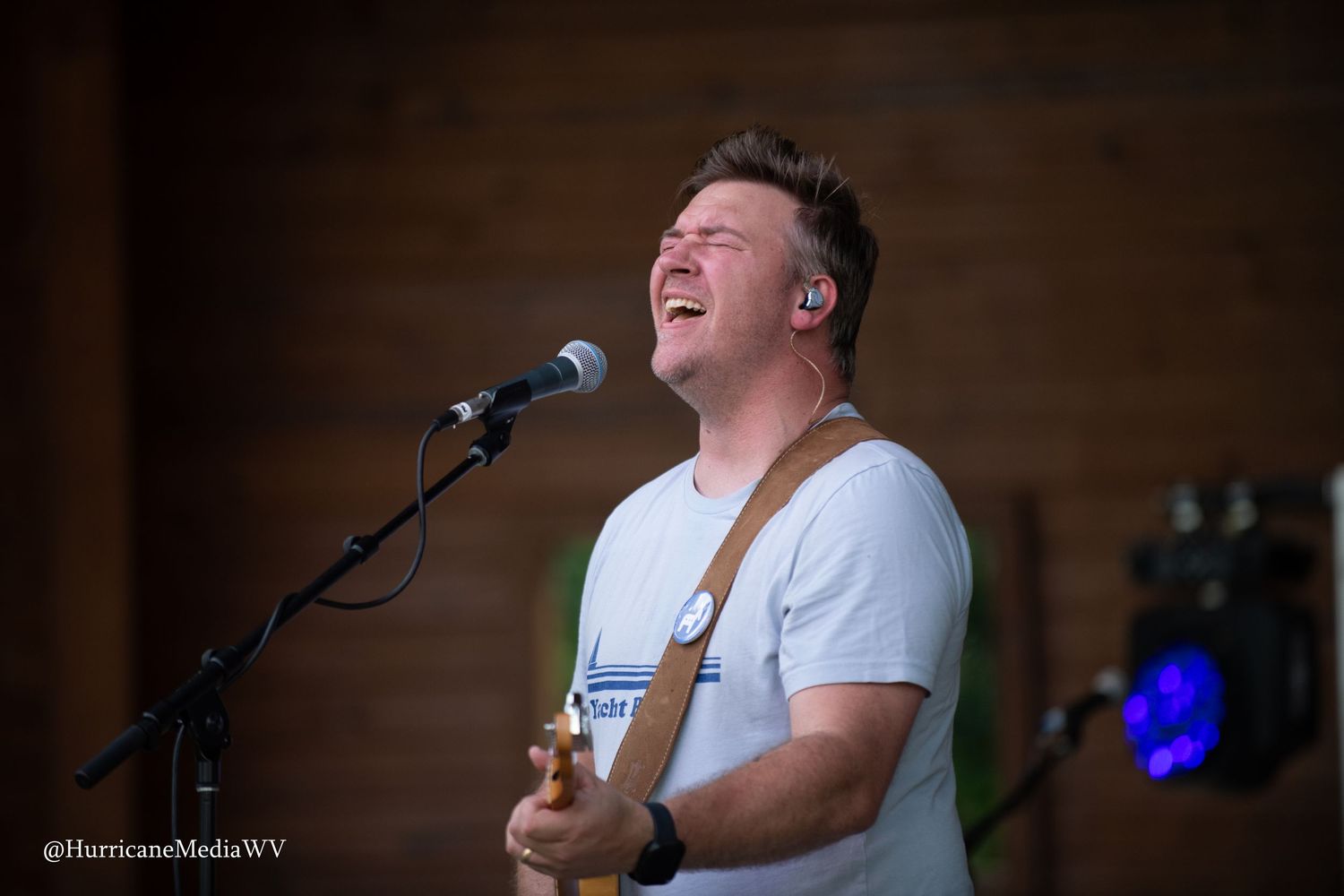 Performer in white t-shirt sings passionately into microphone during live performance with stage lighting effects.