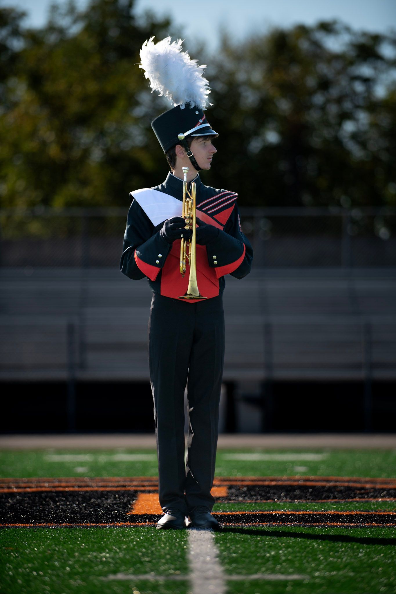 A marching band member in formal uniform stands at attention holding an instrument.