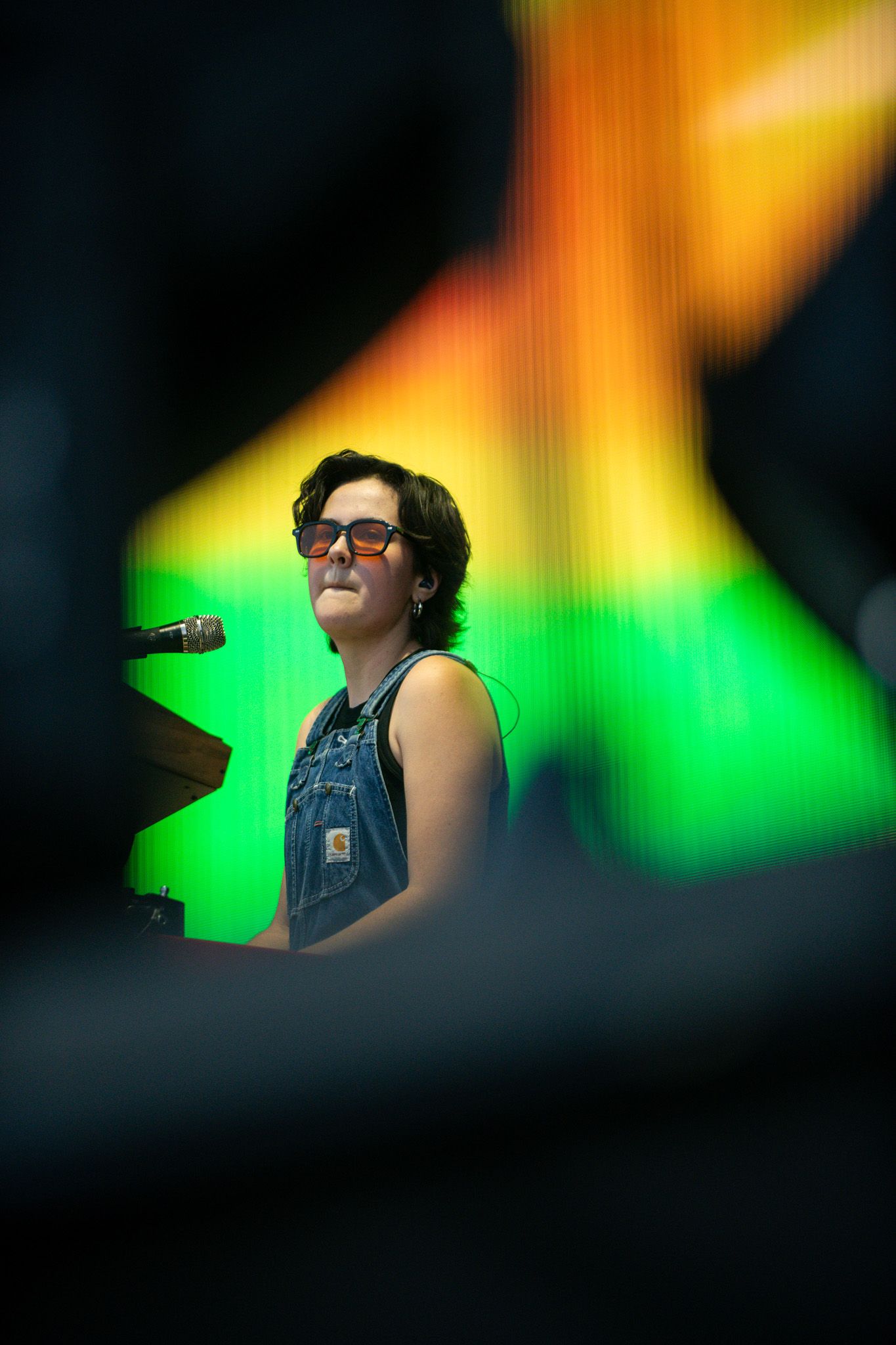 Musician in denim overalls and glasses performs on stage with colorful green and orange lighting effects in background.