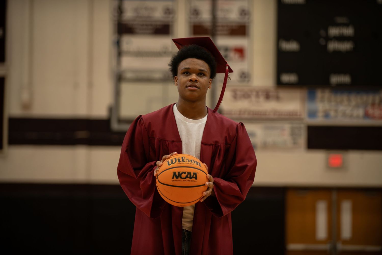 Graduate in maroon cap and gown holds basketball in gymnasium setting.