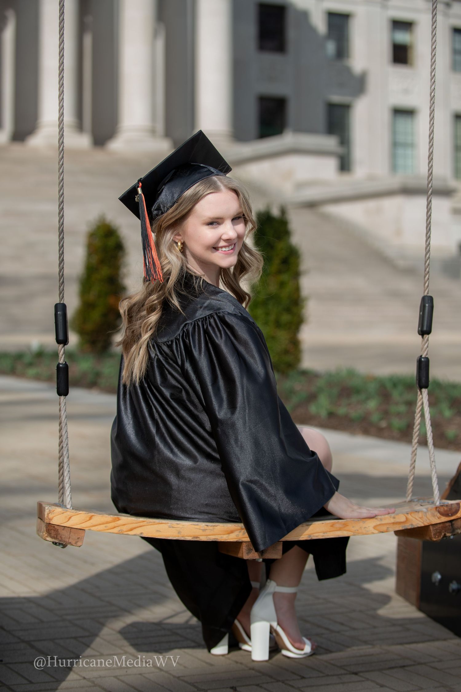 Graduate in black cap and gown sits on wooden swing in front of stone building during graduation photo session.