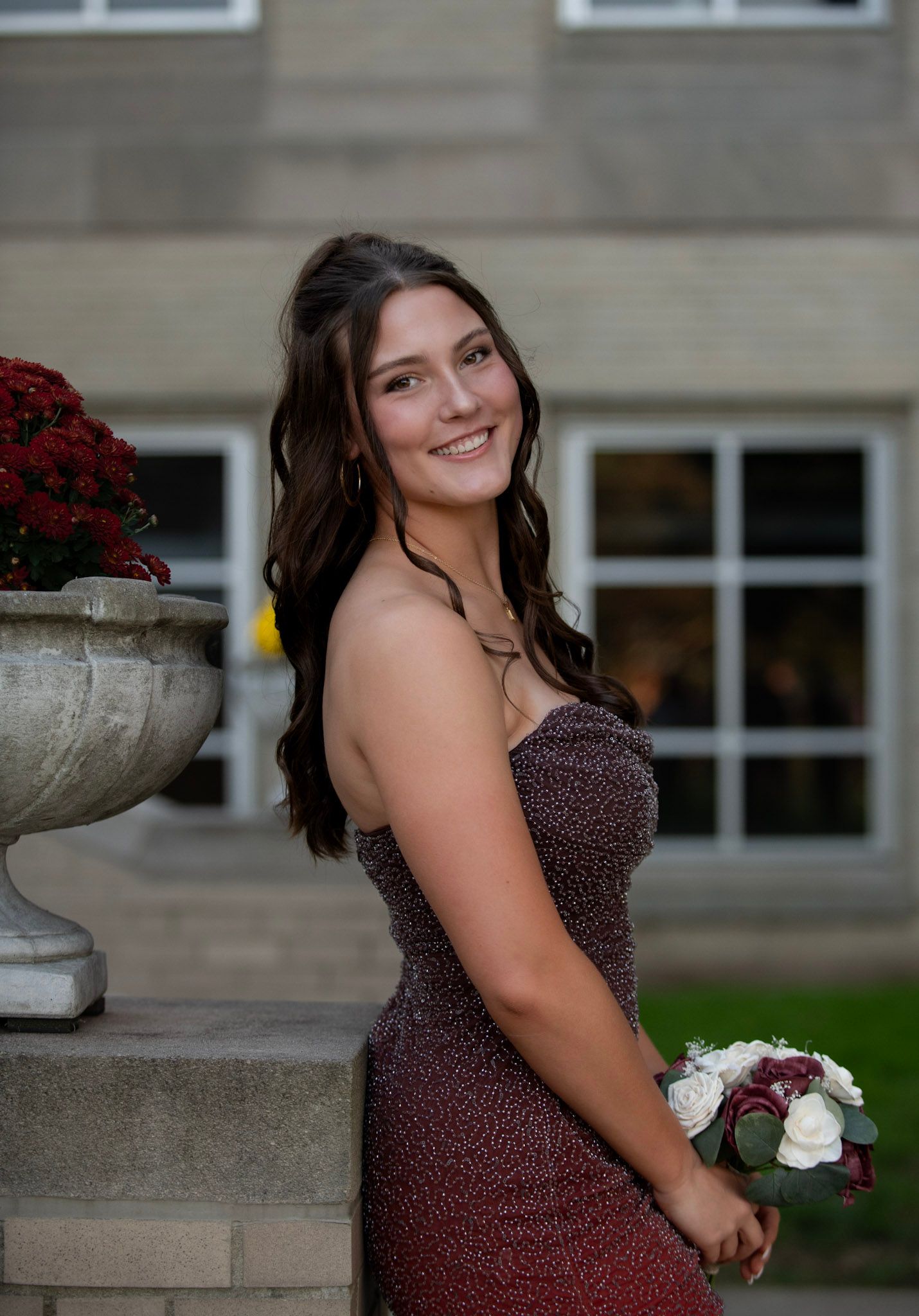 Woman in burgundy formal dress poses next to decorative planter with flowers.